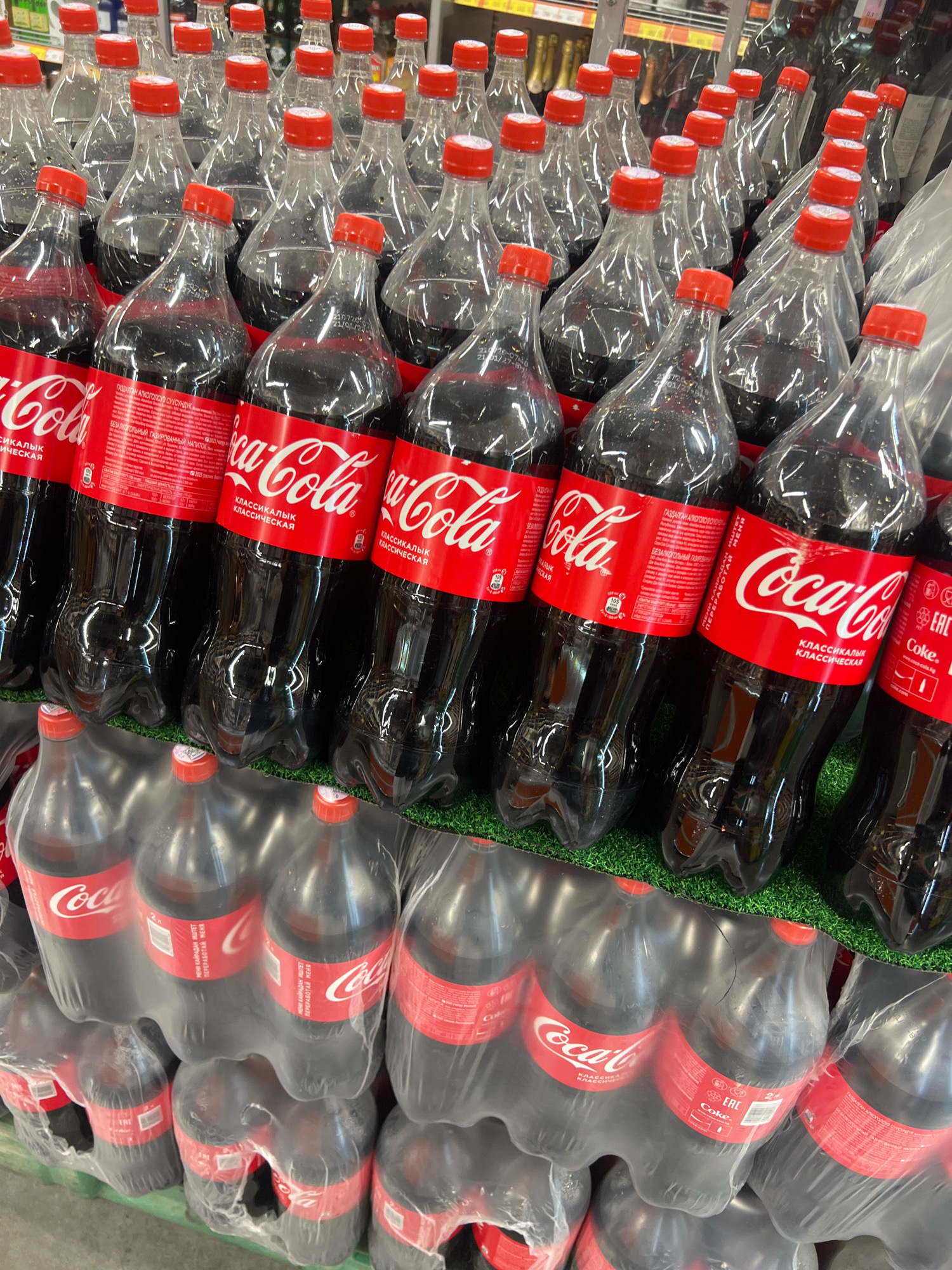 The image shows a close-up, slightly angled view of multiple two-liter bottles of Coca-Cola Classic. The bottles are arranged in rows and are primarily seen from the front, with their iconic red labels facing the camera. The liquid inside the bottles appears dark brown, consistent with Coca-Cola. The bottles are made of clear plastic and have red caps. Some bottles are individually wrapped in clear plastic, while others are grouped in multi-packs also covered in plastic. The bottles are displayed on a green, textured surface, possibly artificial grass or a mat, which is common in retail displays. The background is blurred but suggests a retail environment with shelves and other products, typical of a supermarket or convenience store. No people or specific landmarks of Osh City, Kyrgyzstan are visible in this image; it is a generic retail product display.