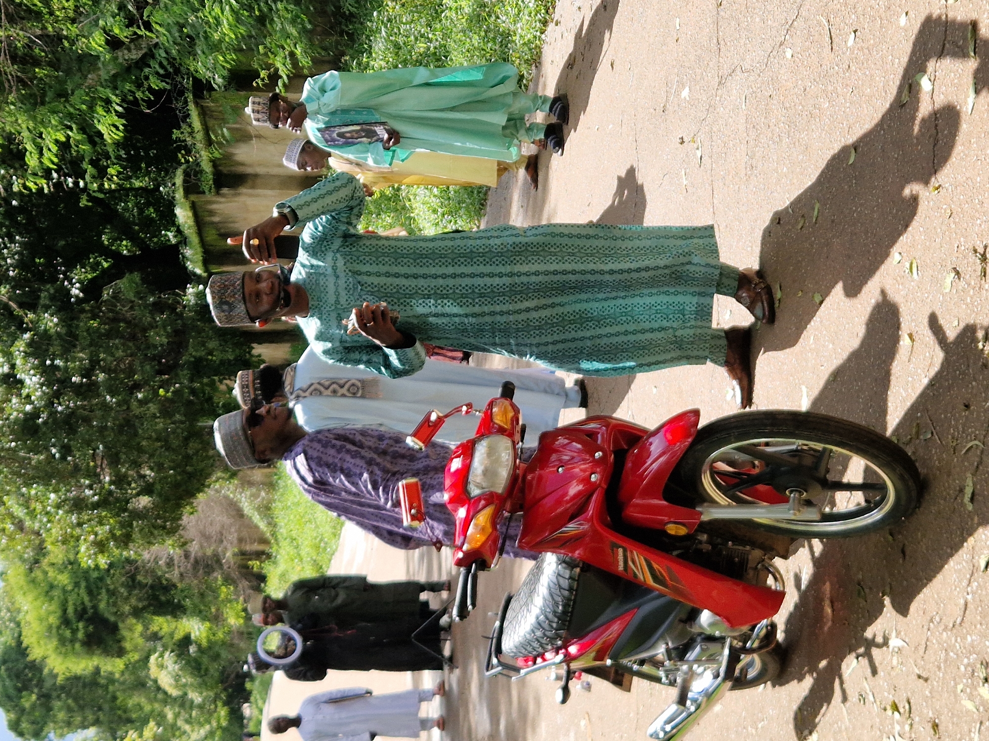 In Jos, Nigeria, on a sunny day, a group of Muslim men are gathered on a paved path beside a wall and lush greenery. A red motorcycle is parked in the foreground, its front wheel and handlebars visible. The men are dressed in traditional attire, including long robes and caps. One man, wearing a light green robe, gestures with his right hand, holding a small object, possibly a phone. Another man to his right, in a light blue robe, holds a framed image. The man in the purple robe on the left appears to be talking with someone not fully in frame. The atmosphere suggests a social gathering or event, possibly religious or ceremonial. The lighting indicates it is midday, casting distinct shadows on the ground.