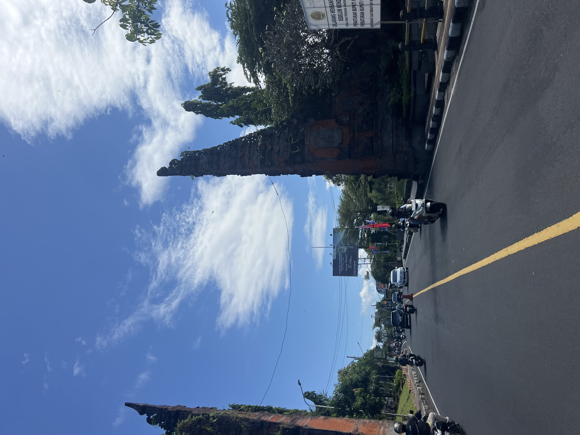 The image shows a street in Denpasar, Indonesia, with a traditional Balinese gate on either side of the road. The sky is blue with scattered white clouds. Traffic is visible on the road, with several motorcycles and a few cars. A yellow line divides the road. A sign with Indonesian text is posted near one of the gates. Trees and foliage are present along the roadside. An advertising billboard is also present on the side of the road.