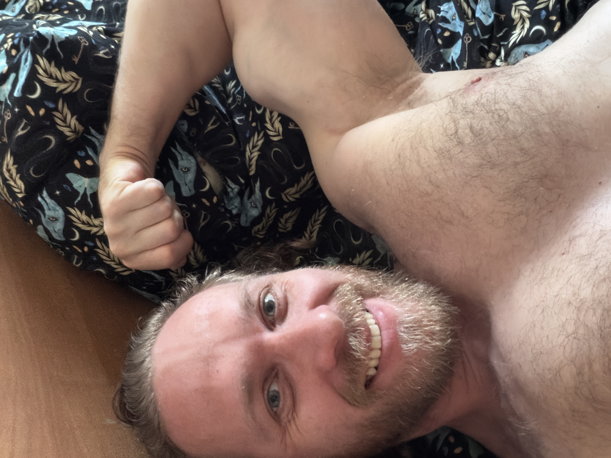 A topless man is lying on his back on a bed with a dark blue and black patterned sheet. He is smiling at the camera with his eyes open and mouth slightly ajar, showing his teeth. He has a beard and light hair. His left arm is bent and raised with his fist clenched. The background appears to be a bedroom. There is no indication of the location being in Saint Petersburg, Russia.