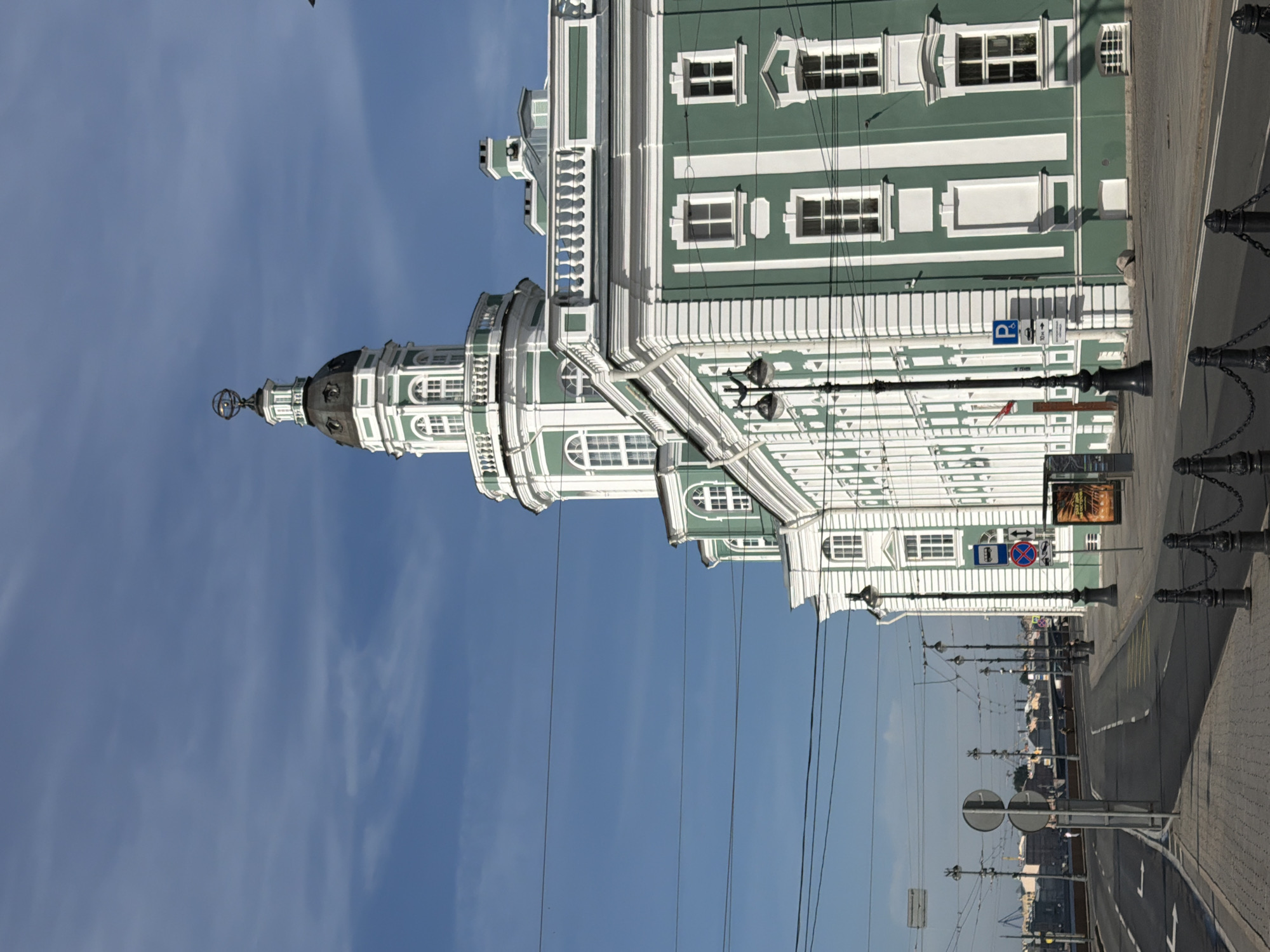 The image depicts the Kunstkamera museum in Saint Petersburg, Russia, on a clear day with a blue sky and scattered clouds. The grand, historic building, painted in a distinctive green with white trim, features an imposing central tower topped with an ornate globe. The foreground shows a street scene with a paved sidewalk, a road with lane markings, and several streetlights. Traffic signs and a bus stop are visible, along with black metal bollards and chains lining the sidewalk. In the background, a bridge and other buildings are visible along the waterfront, suggesting the museum's location on the Neva River. There are no people clearly visible in the image.