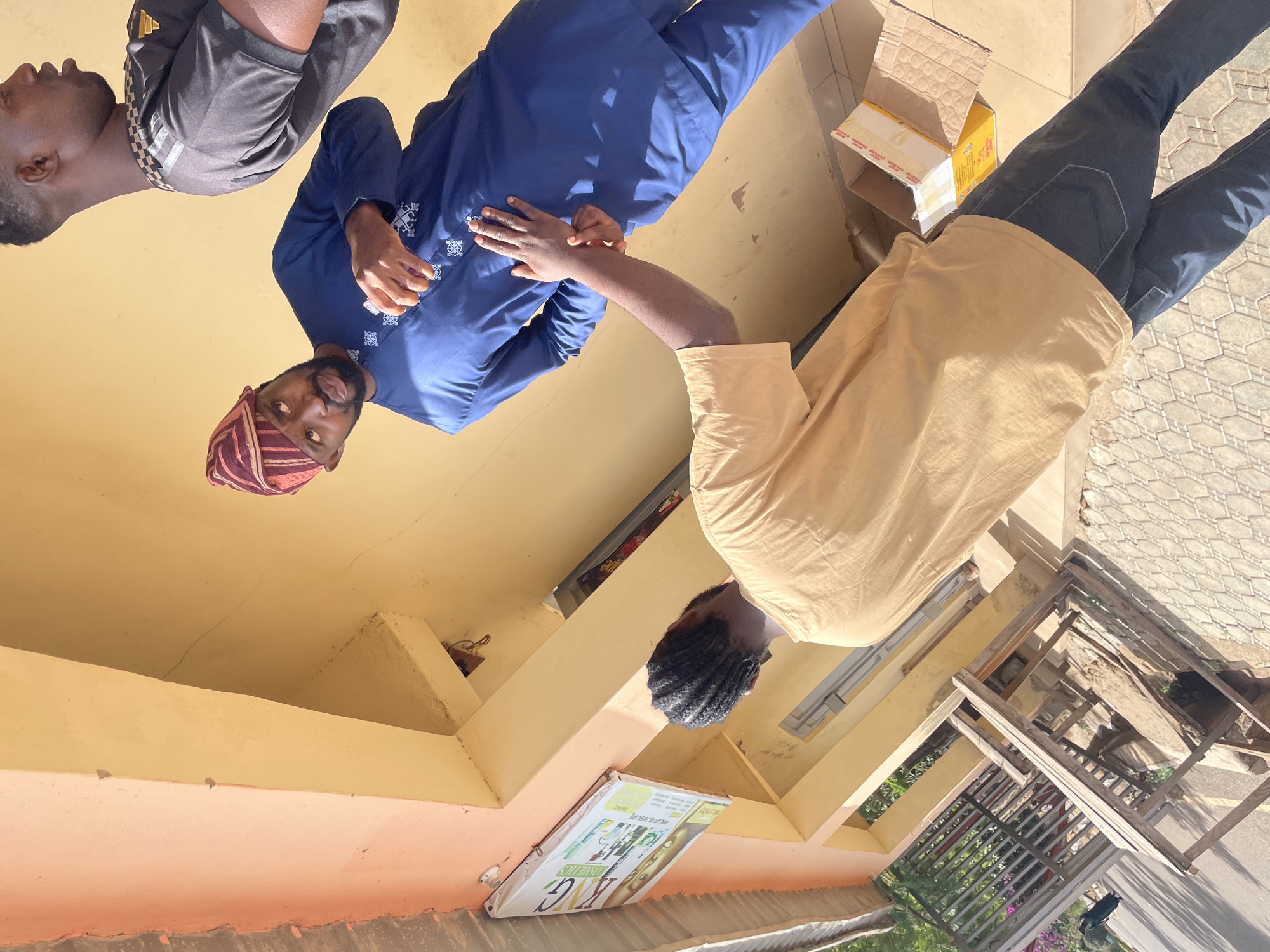 The image captures three men engaged in an interaction outdoors during bright daylight, likely in Jos, Nigeria. The setting features a building with walls painted in warm tones of yellow and peach/orange, and a patterned tiled or paved floor on the right.
On the left, a man in a grey short-sleeved top with black and white checkered detailing stands looking towards the center. In the middle, another man wears a blue long-sleeved garment, possibly a traditional kaftan or dashiki, adorned with white embroidery. He also wears a striped, multi-colored (reddish-brown and cream) cap or hat, commonly seen in West African attire. He has a beard and is extending his right arm towards the third man.
On the right, a third man, with short dark braided hair, is dressed in a light tan/beige long-sleeved shirt and dark trousers. He extends his left arm towards the man in blue, suggesting a handshake, a gesture of interaction, or an exchange. The overall scene appears to be a casual social encounter or a meeting.
In the background, on the far right, there is a wooden frame structure, possibly a stand or scaffolding. Beyond this, green foliage and purple flowers are visible. Below the man in tan, against the building's lower wall, a white sign or poster is displayed, prominently featuring "KING COSMETICS" in white letters, along with the number "200" and other smaller, less legible text like "DELICIOUS." Near the man in tan, two cardboard boxes are visible on the ground; one is open, and the other is sealed, bearing text such as "DELICIOUS BISCUITS" and "BEST QUALITY BISCUITS." The clear, bright lighting and visible shadows indicate it is daytime, likely mid-morning to afternoon.