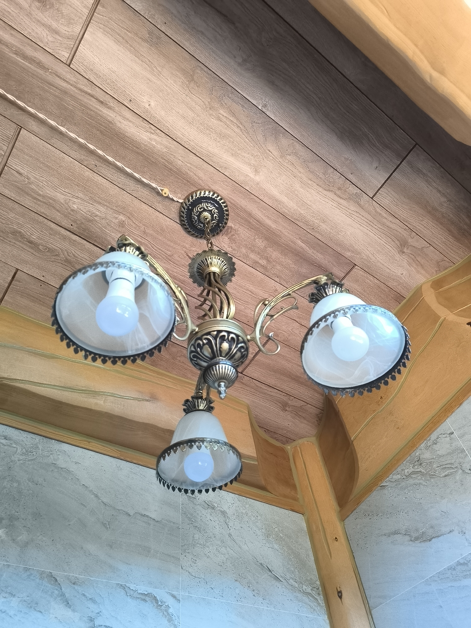 A brass chandelier with three glass shades hangs from a wooden ceiling. Each shade contains a white LED light bulb. The chandelier is ornate with decorative metalwork. The ceiling is made of wooden planks. To the right and below the chandelier, there is a decorative wooden beam. The wall below the beam appears to be tiled with a gray, marble-like pattern. The image was taken indoors, likely in a room with a rustic or traditional architectural style. There are no people or animals visible in the image. The location is identified as Malyy Baklaniy, Russia.