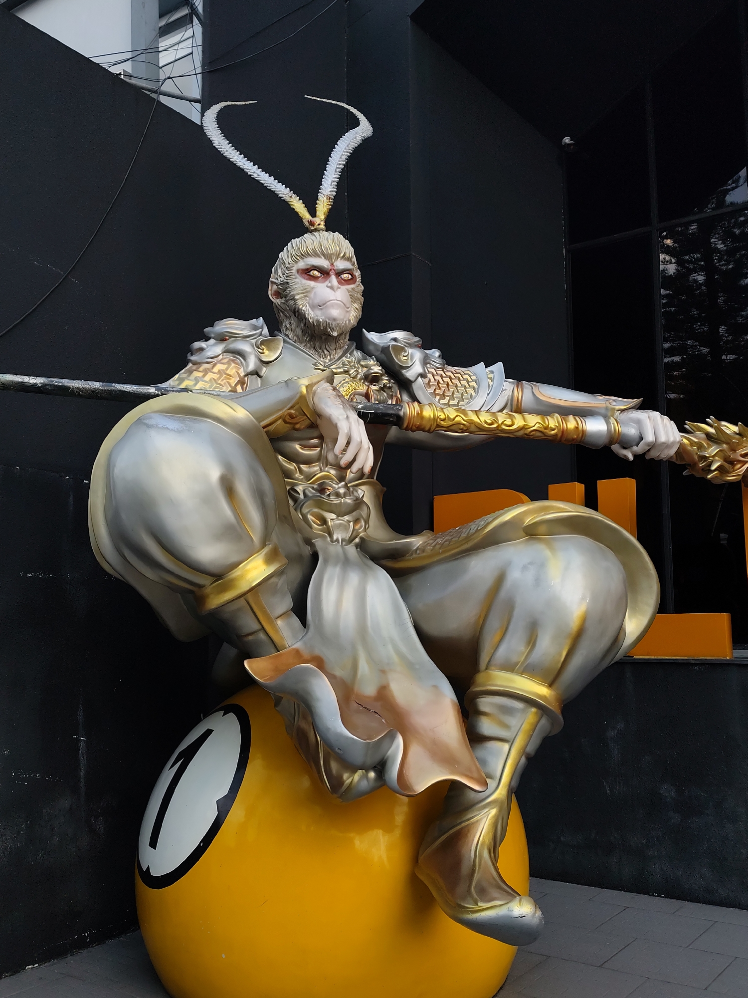 A statue of Sun Wukong, also known as the Monkey King, sits atop a large yellow billiard ball with the number 1 painted on it. The statue is positioned in front of a building with a black exterior wall. Wukong is depicted in silver and gold armor, holding a staff. The statue appears to be located outside of a business, with a glimpse of an orange logo visible behind the figure. The location is Da Lat, Vietnam.
