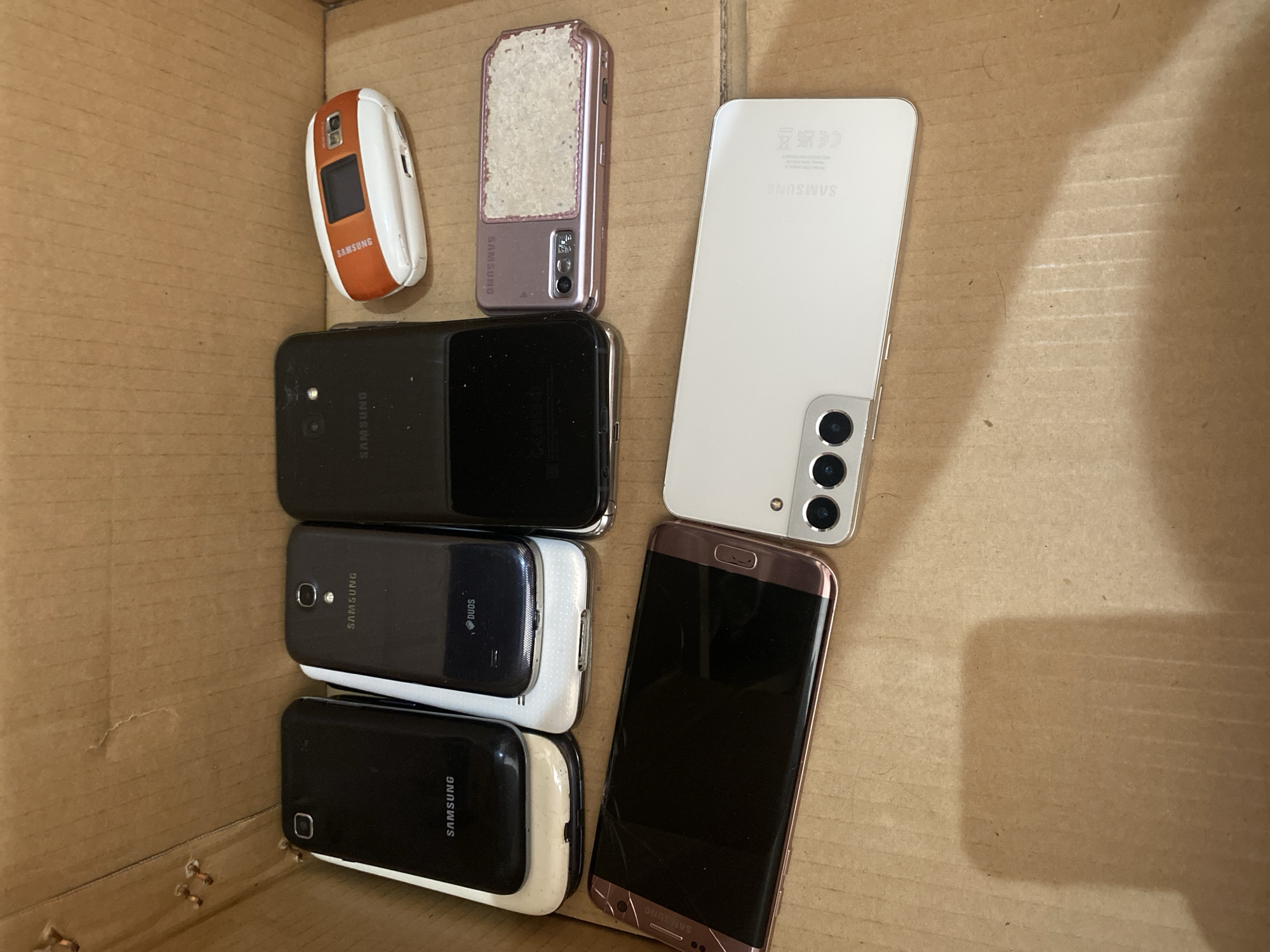 A collection of Samsung mobile phones is neatly arranged in a cardboard box. The phones are of various models and colors. In the foreground, a rose-gold colored smartphone with a curved screen is positioned to the left. To its right, a white smartphone with a triple camera array is visible. Behind these, a series of older Samsung phones are stacked. A small, orange and white flip phone is situated at the top right of the arrangement. A rose-gold colored flip phone with a textured panel is next to the black smartphone. The background consists of the textured surface of a cardboard box, which shows signs of wear. The lighting is dim, casting shadows on the cardboard surface, suggesting an indoor environment. There is no indication of the specific location within Chișinău, Moldova, or any people present in the scene.
