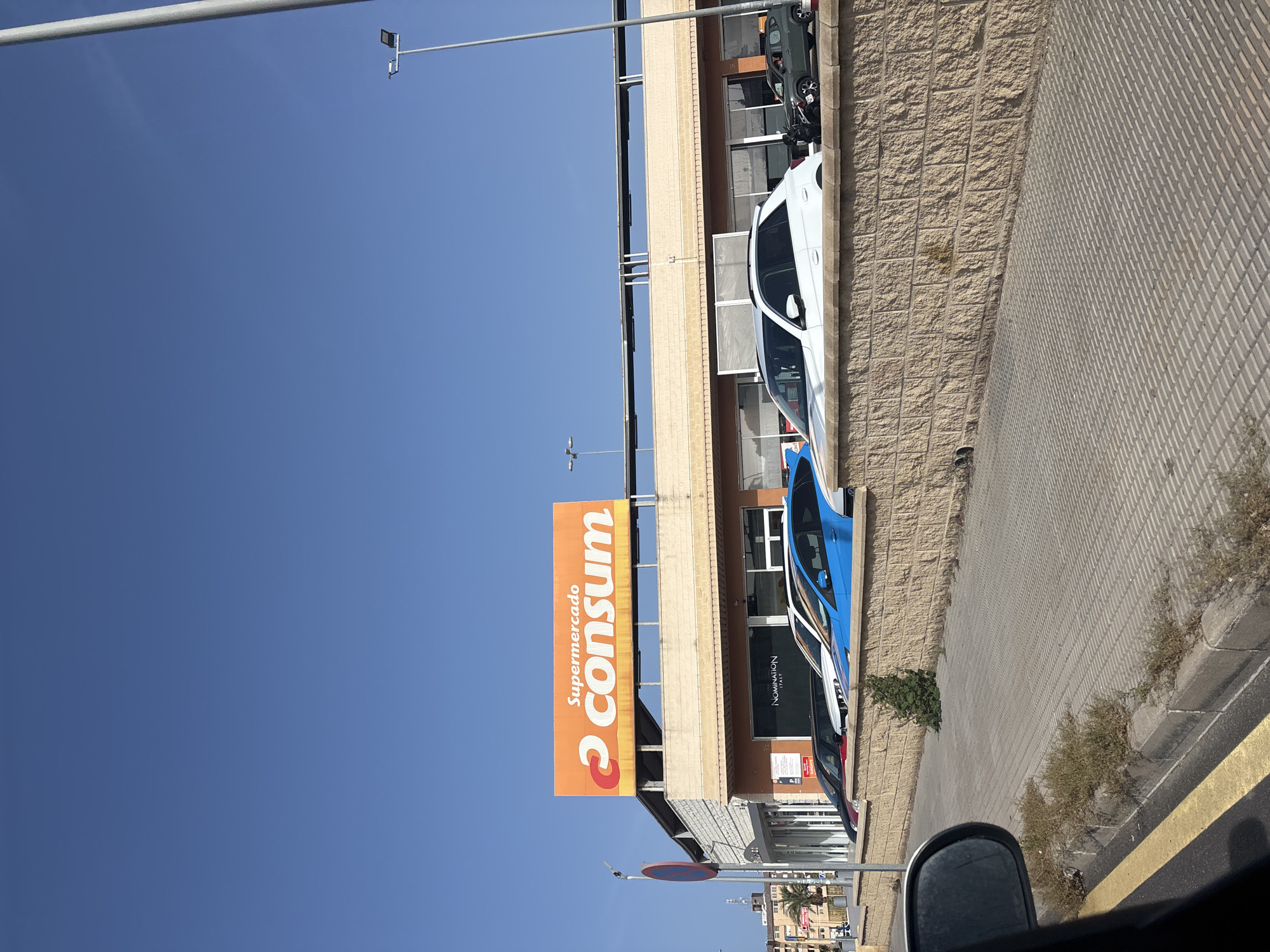 This image shows a street view in Orihuela, Spain, with a Consum supermarket visible. A large orange sign with the Consum logo and "Supermercado" written above it is mounted on the building. In front of the supermarket, several cars are parked: a dark colored car, a red car, a white car, and a blue car. A textured brick wall runs along the sidewalk in front of the cars. The sidewalk is paved with rectangular tiles. A street lamp and a traffic sign are also visible. The sky is clear and blue. The image appears to have been taken from a vehicle, as a car's side mirror is visible in the bottom left corner.