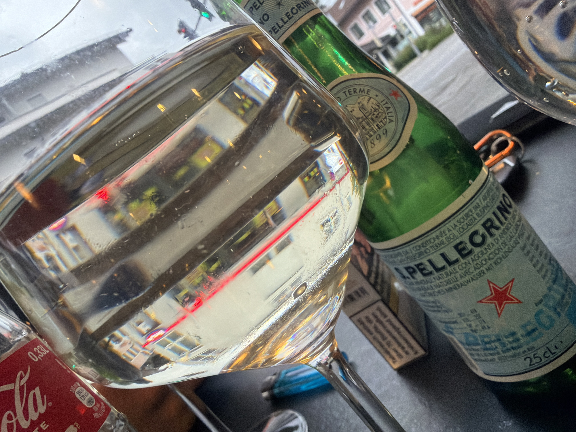 Here is a description of the image:
The image shows a close-up view of a table setting, likely in a café or restaurant. Prominently featured is a wine glass filled with a clear liquid, possibly white wine or sparkling water. Next to the glass is a green glass bottle of San Pellegrino sparkling water, with its label visible. Part of a Coca-Cola bottle is also visible in the bottom left corner.
Reflections in the glass provide a distorted view of the surroundings, including buildings and what appears to be a traffic light. The location appears to be in an urban setting. Based on the buildings and the general atmosphere, the location could be in a city in Europe. The city in the image appears to be Garmisch-Partenkirchen, Germany. Other items visible on the table include a cigarette pack and an unidentified object with a ring. The table itself has a dark surface.
