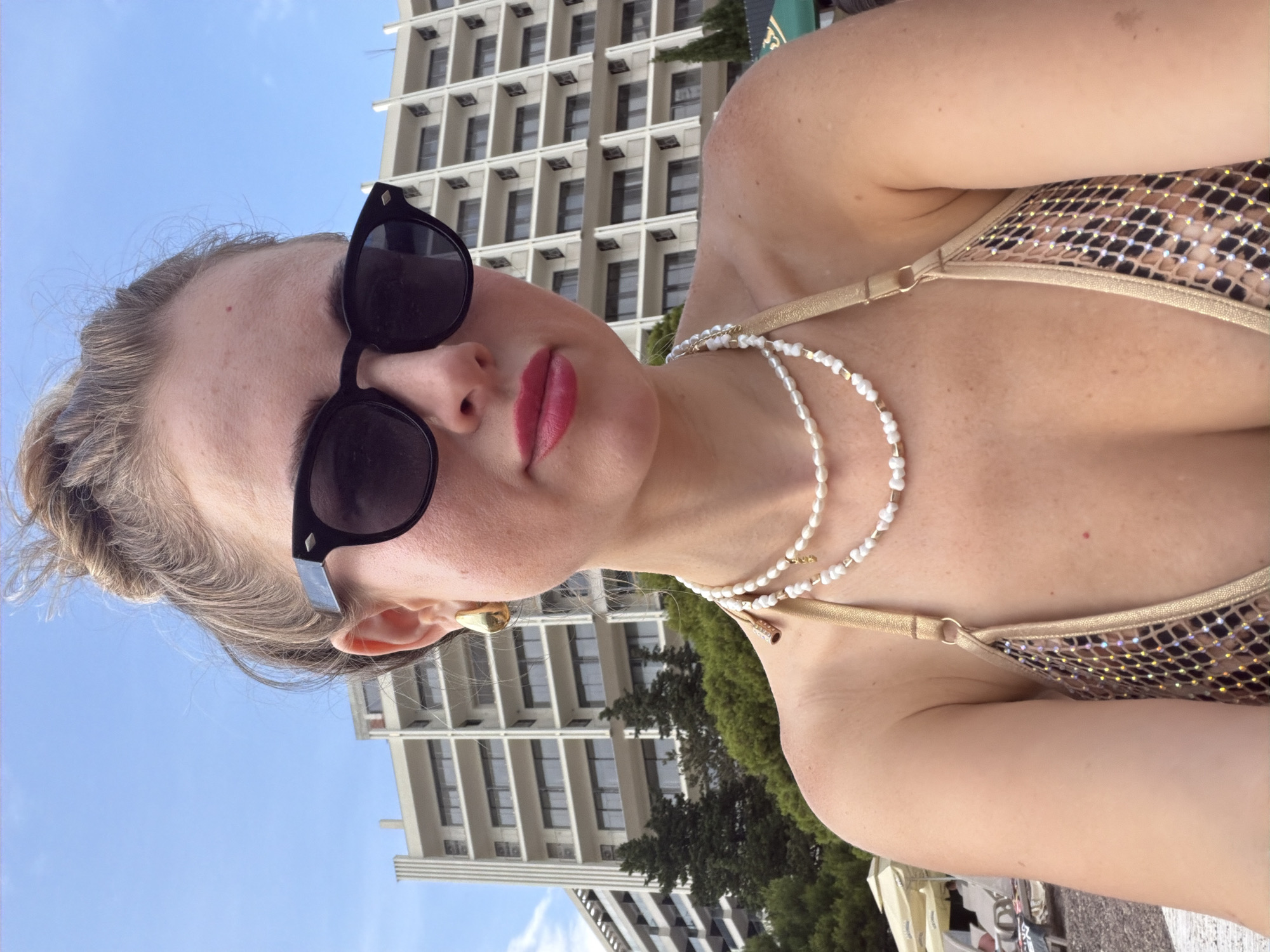 The image shows a woman wearing sunglasses and a bikini, with a building and trees in the background. She is looking up and to the right, with her lips slightly parted. The sun appears to be shining, and the sky is blue. The woman is wearing a pearl necklace and a gold earring. The bikini top is gold with a leopard print pattern. The building in the background is tall and modern, with many windows. There are also green trees and a clear blue sky visible. The scene appears to be set outdoors, possibly at a beach or resort.