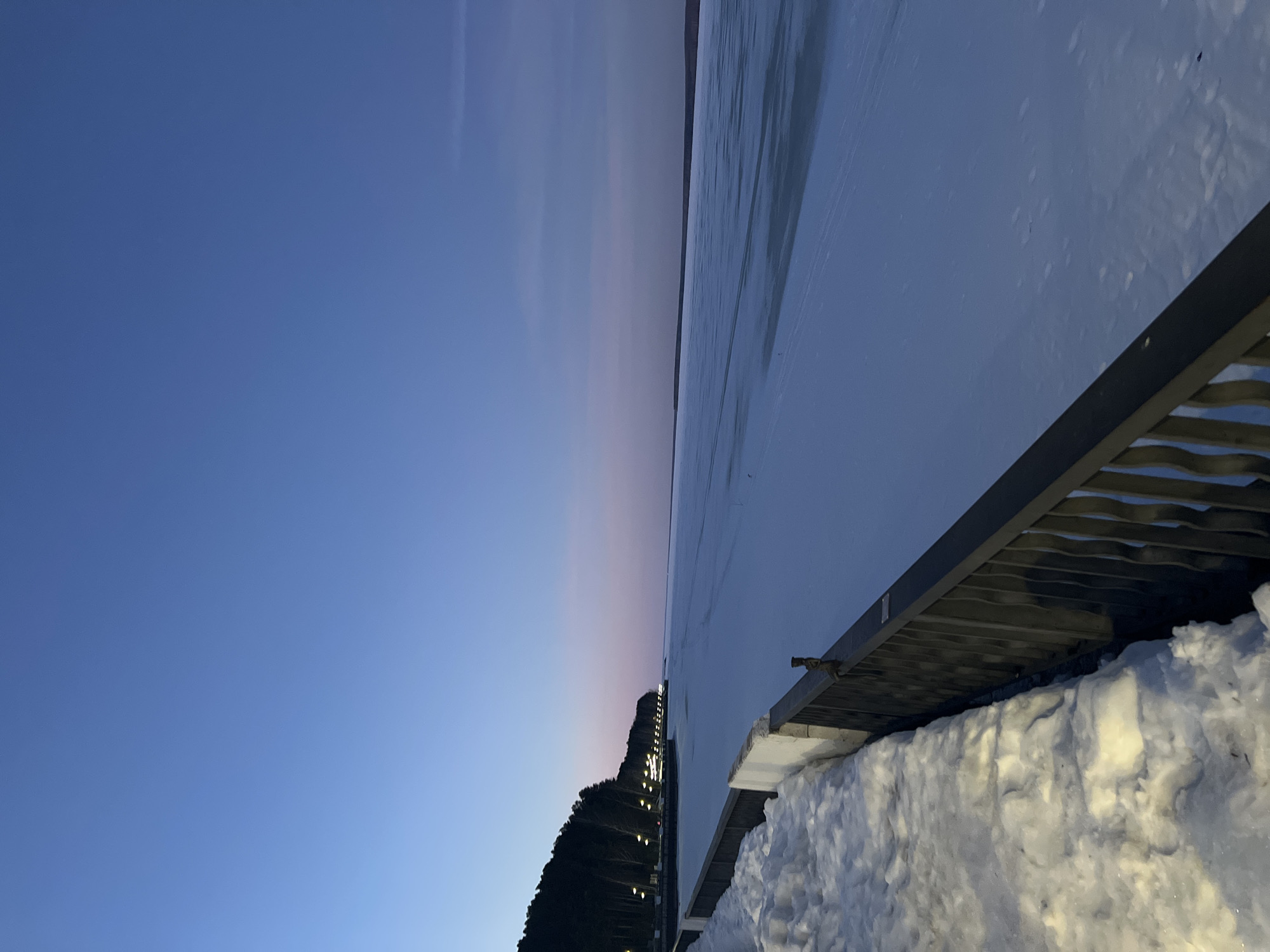 The image captures a winter twilight or evening scene over a vast frozen body of water, likely the Kama River or Votkinsk Reservoir, adjacent to the city of Chaikovsky, Russia. In the immediate foreground, a dark metal railing with vertical bars extends diagonally from the lower right toward the center-left. Adjacent to the railing on the left is a significant accumulation of lumpy, partially snow-covered ground, which transitions into a concrete barrier visible beneath the railing. Beyond the railing, the expansive surface of a frozen body of water stretches into the distance, appearing as a mix of white snow cover and patches of bluish-grey ice. Along the left bank of the frozen water, a dense line of dark coniferous trees is visible, below which a series of evenly spaced, glowing yellow-orange streetlights illuminate a promenade or embankment. The sky above presents a clear gradient, transitioning from a dark blue overhead to a lighter purplish-pink hue near the horizon. No individuals, vehicles, or animals are discernible within the frame.
