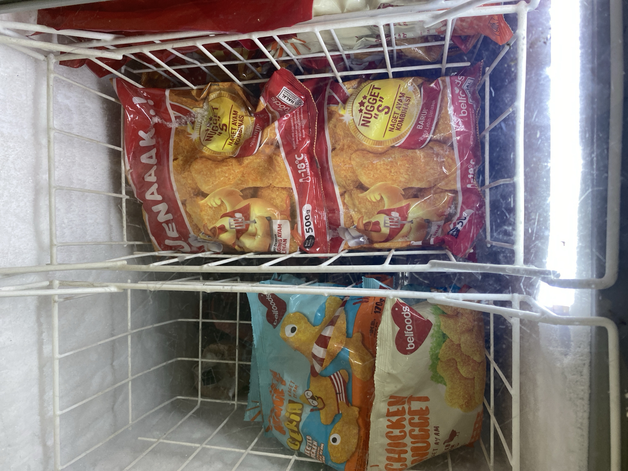 The image shows a close-up view of a freezer chest filled with various frozen food products, primarily chicken nuggets. The freezer is divided by white metal racks, separating different sections of the products. On the left, there are at least two bags of "Chicken Nugget" from the brand "belfoods," one with a light blue packaging featuring cartoon characters and the other with white packaging showing an image of nuggets. To the right, several red and gold bags of "NUGGET S" are stacked, also from "belfoods," with images of fried chicken nuggets and text indicating "NAGET AYAM KOMBINASI" and "Rasa Lebih Lenaaak." Some bags also display "HALAL" certification. The background shows the frosted interior of the freezer. There are no people or animals visible in the image. The scene is indoors, within a commercial freezer unit, suggesting a retail or food service environment. The location is identified as Payatieng, Indonesia, which implies a commercial establishment such as a convenience store, minimarket, or restaurant in that area.