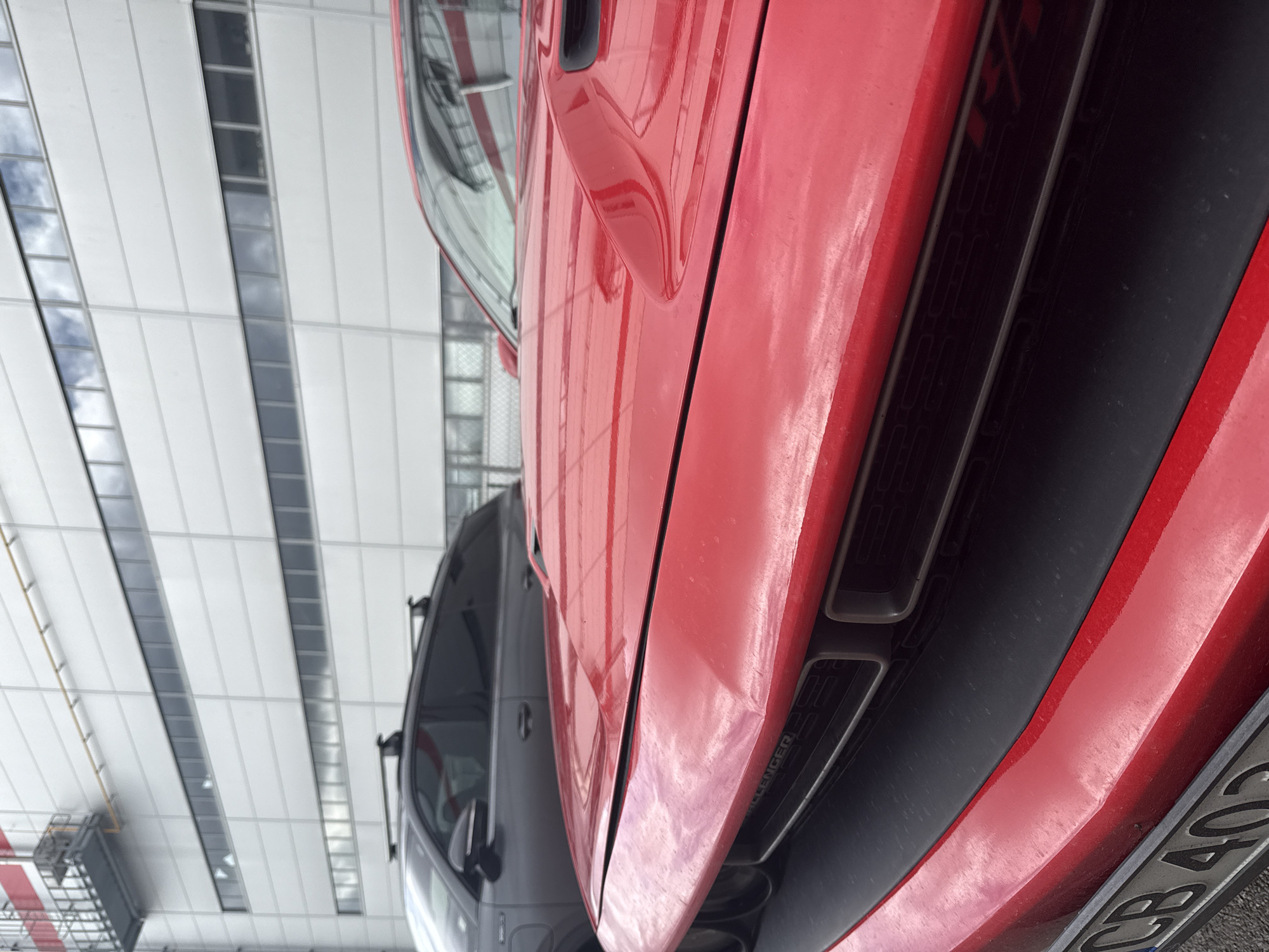 The image shows the front end of a red Dodge Challenger parked indoors, likely in an industrial or commercial building based on the visible interior architecture (white walls, overhead lighting and structural supports). Another dark colored car is visible in the background, parked near the Challenger. The Challenger's license plate starts with "CB 402", which indicates it is registered in Sofia, Bulgaria. The image focuses on the front of the red vehicle, specifically the front grill, and the car's body.