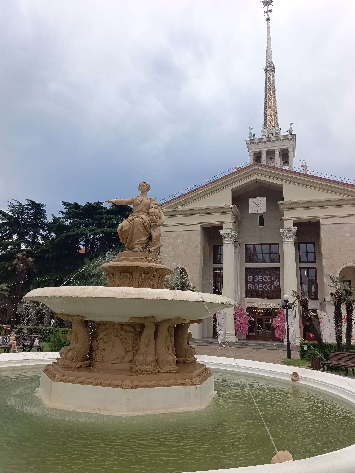 The image shows a fountain in front of a large building with a prominent spire. This is the Sochi Sea Port, a landmark in the city of Sochi, Russia. The fountain is ornate, with a statue of a woman and child at its center, and its base decorated with figures of fish. Water is spraying from the fountain into the pool. The building behind the fountain is a grand structure with classical columns and a large clock on its facade. Banners with the word "BOSCO" are visible on the building. Lush green trees frame the scene on the left, and palm trees are present on the right. The sky is overcast. Several people are visible in the background, walking and sitting on benches.