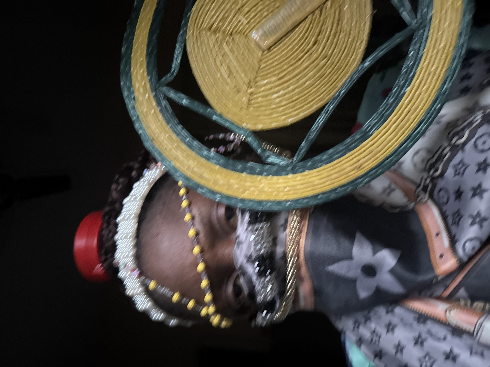 A medium shot shows a person wearing an elaborate headpiece composed of braided hair, beads, and a red cylindrical element, placed beneath a woven, circular object featuring yellow and green hues. The individual is also adorned with necklaces, and is draped in a scarf or garment patterned with floral and star motifs. The background is dark. The location is unknown.