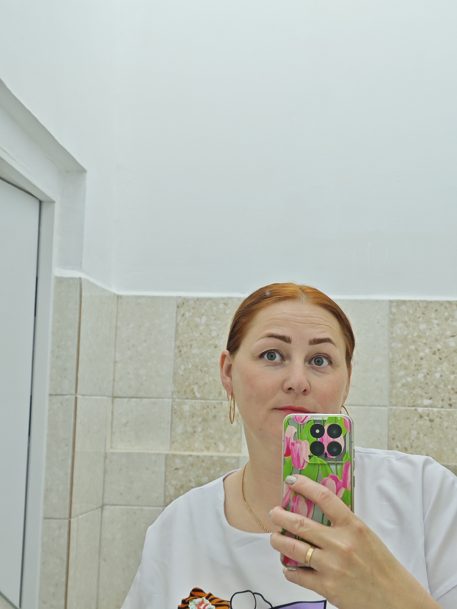 A woman with reddish-brown hair is taking a selfie with a mobile phone in a bathroom. The phone has a clear case adorned with a floral design of pink tulips and green leaves. She is wearing gold hoop earrings, a gold chain, and a wedding ring on her left hand. She is wearing a white t-shirt, and the background consists of beige tiled walls and a white ceiling. A white door frame is visible in the left side of the frame. It is not possible to determine if she is in Фоки, Russia, based solely on this image.