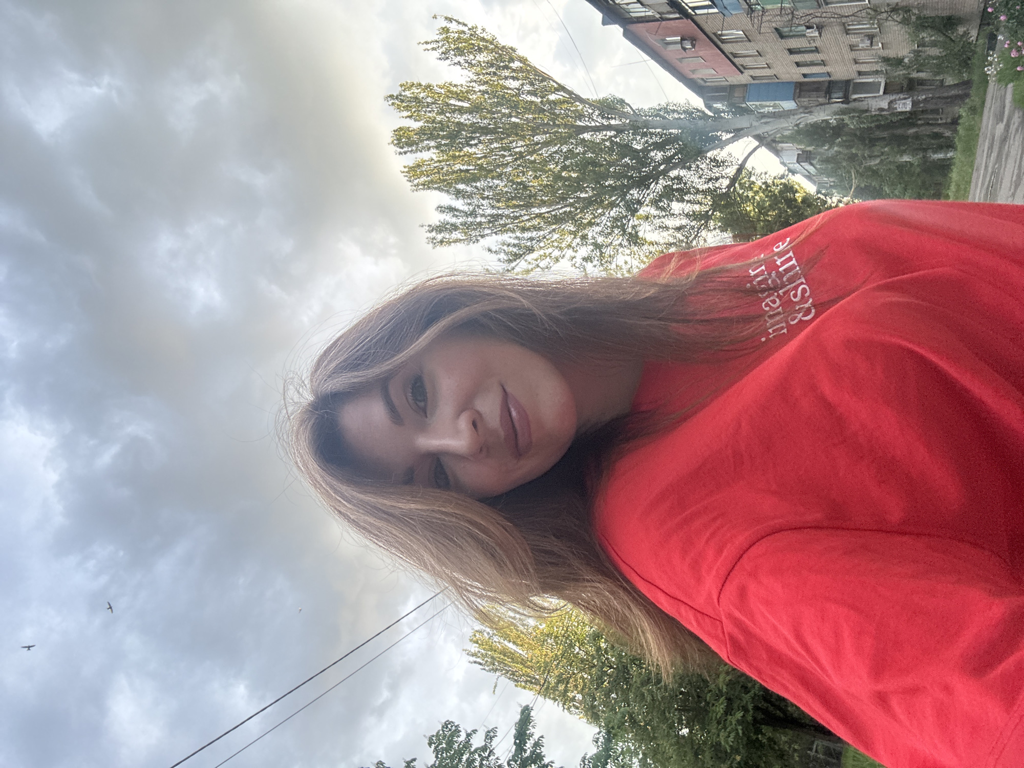 A fair-skinned woman with medium-length blonde hair is pictured in a casual, eye-level selfie. She is wearing a red t-shirt. Her face is turned towards the camera with a slight smile.
In the background, the sky is overcast with grey clouds. There are trees and an urban landscape visible. The building architecture is typical of post-Soviet urban development. The precise location in Kryvyi Rih, Ukraine, is difficult to determine without more specific landmarks. The shot is taken from below looking up. There is a bird flying in the sky. Also, there are wires hanging above the trees and buildings.