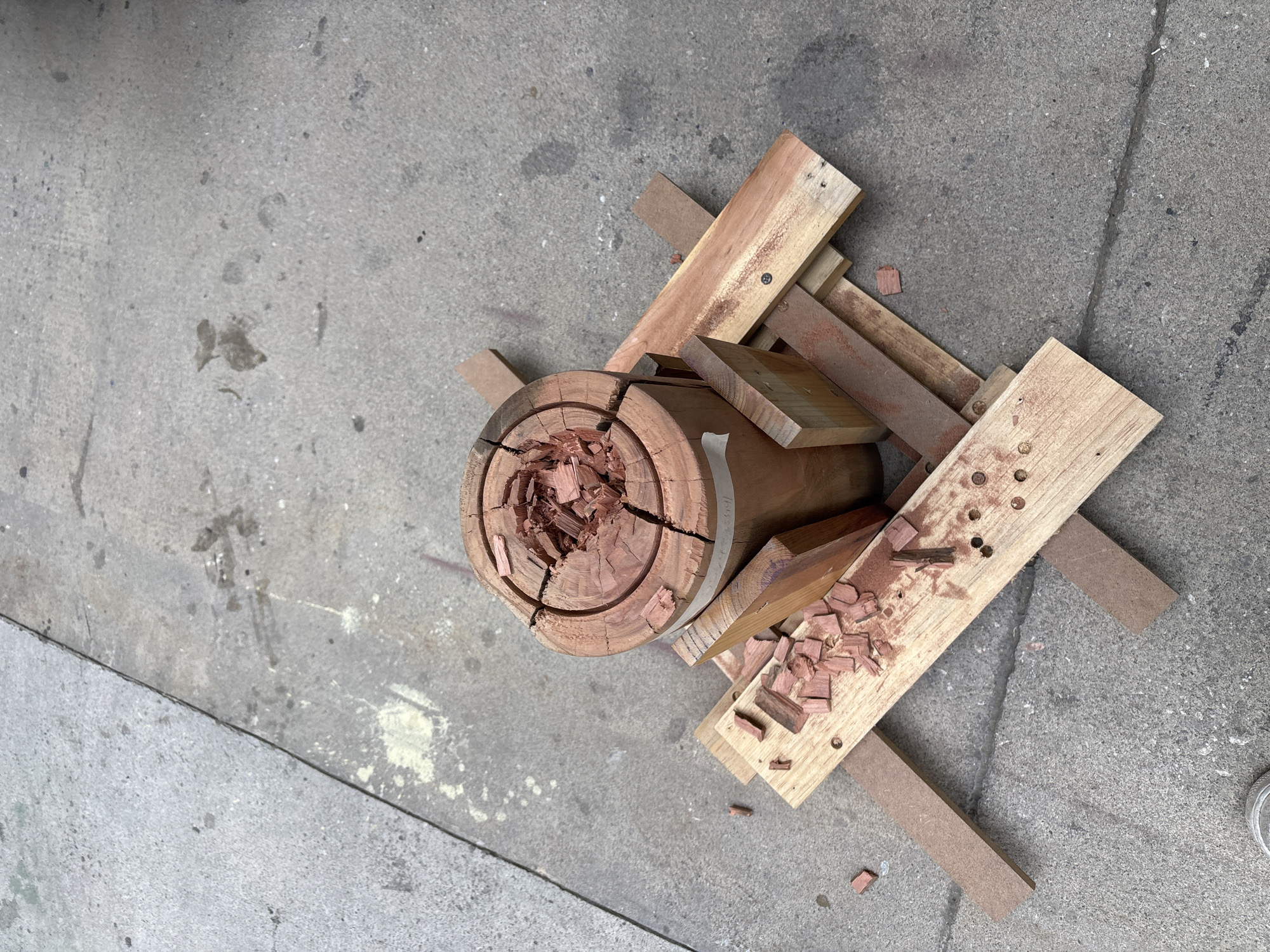 An overhead shot displays an assortment of woodworking materials and debris laid out on a light grey concrete surface, possibly a sidewalk or outdoor work area. The central object is a cylindrical log or stump of reddish-brown wood, heavily cracked on its top surface, revealing distinct concentric growth rings. Its core is deeply carved or fragmented, filled with numerous small, reddish-brown wood chips and shavings, indicating recent manual work. A narrow metallic band is partially visible encircling the log.
To the right of the log, a cluster of various wooden planks and blocks are arranged. Some pieces are lighter in color, while others match the reddish tone of the log. Several planks feature multiple drilled holes or screw indentations. Interspersed are smaller, uniform blocks of what appears to be engineered wood, like MDF. Reddish wood chips and sawdust are scattered across these wooden pieces and onto the surrounding concrete.
The concrete ground shows a rough texture, mottled with various stains, dirt, and some pale yellow powdery residue. Faint cracks and lines crisscross the surface. The scene is brightly lit, suggesting daytime. No people are visible, nor is any text. The arrangement strongly implies an ongoing or recently completed woodworking, carving, or craft activity. While the setting is outdoor on concrete, no specific visual cues link it directly to Mexico City.