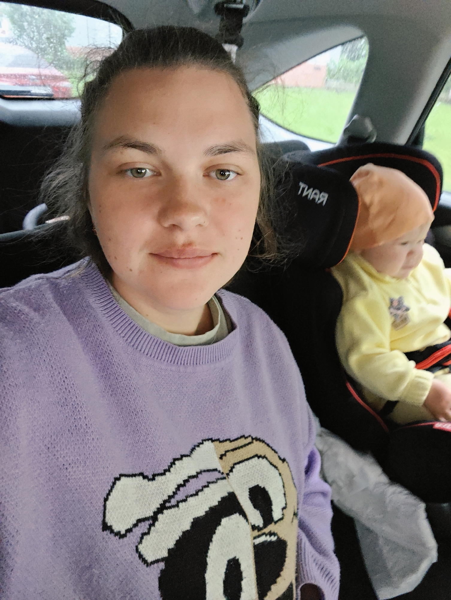 A woman and a baby are in a car, likely in the backseat. The woman is taking a selfie. She has fair skin, green eyes, and her hair is pulled back. She is wearing a lilac sweater with a cartoon character on it. The baby is seated in a black car seat, wearing a yellow outfit and a light orange hat. The car's headrest is visible behind the baby, with the word "TIAR" printed backward on it. Outside the car window, there appears to be green vegetation and a building. The image metadata indicates the location as Chaikovsky, Russia. The photo appears to be taken during the day, possibly on a cloudy or rainy day.