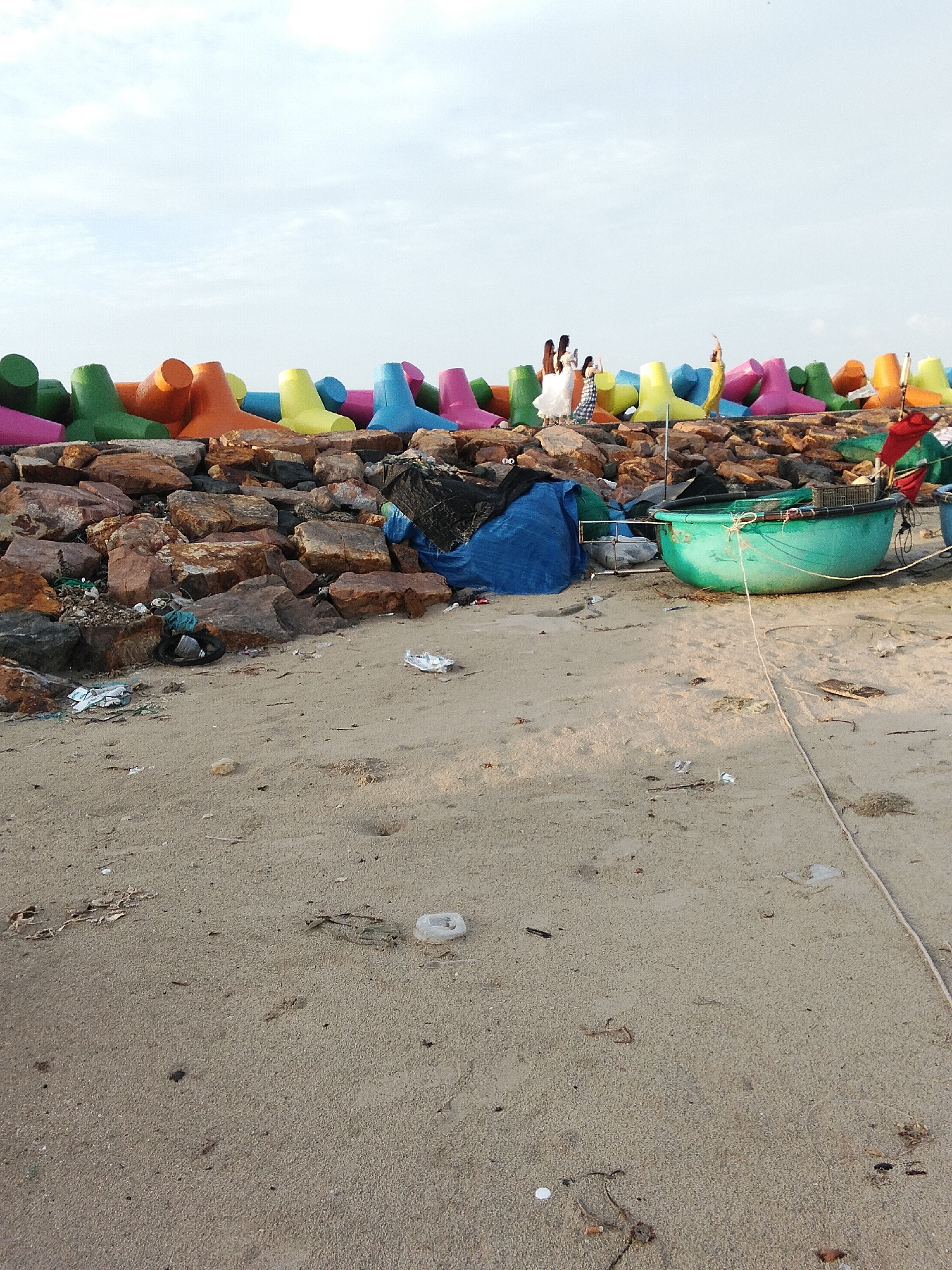 The image depicts a rocky breakwater on a sandy beach, likely in an unspecified coastal area of Vietnam. The breakwater is lined with brightly colored tetrapods, varying in shades of pink, green, orange, yellow, and blue. Some people are visible standing on the rocks near the tetrapods, appearing to be taking photos. One person is wearing a yellow garment and has their arm raised. Below the tetrapods and rocks, a blue tarp and a black tarp are visible, along with a green boat on the sand. The beach itself is sandy and shows signs of debris, including plastic waste and scattered natural materials. A rope extends across the foreground of the image, from the boat towards the bottom right. The sky is overcast with a light haze.