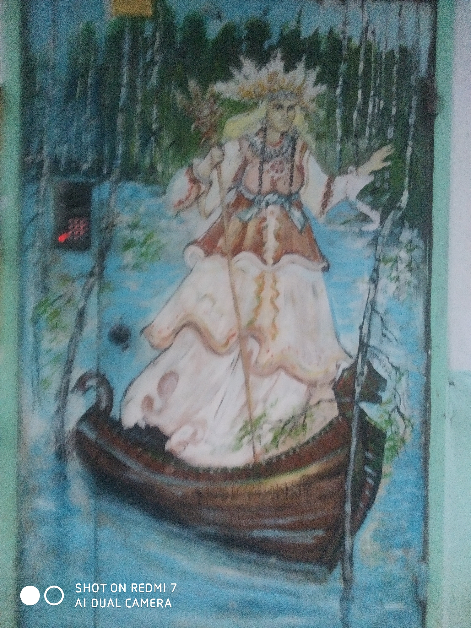 The image shows a painting of a woman standing in a boat on a body of water, possibly a river or lake. The woman is dressed in traditional clothing and has a headdress adorned with white flowers or feathers. She holds a staff in her right hand. The boat appears to be a longboat with a carved prow. The background depicts a forest of birch trees and a blue sky. A dark, rectangular object is visible in the upper left corner, possibly a door panel. The picture was taken on a Redmi 7 AI Dual Camera. The painting is located in the city of Chaikovsky, Russia.