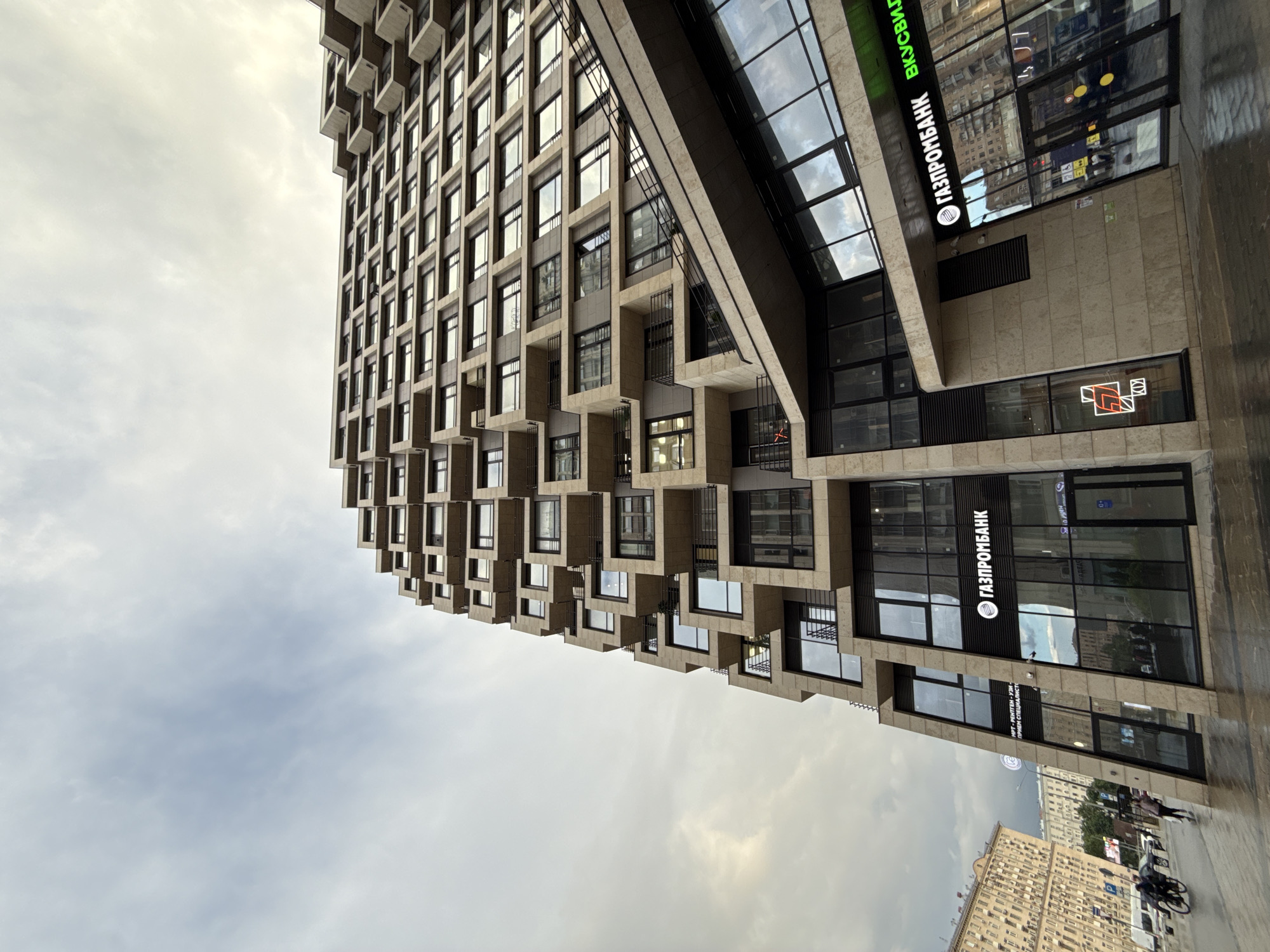 The image presents a low-angle view of a modern building in Moscow, Russia. The building features a unique architectural design with staggered, box-like projections extending outward from the facade. The facade is clad in a light-colored material, possibly concrete or stone, with dark-framed windows.
The lower portion of the building appears to house commercial establishments, including a branch of "Gazprombank," indicated by signage in Russian. Another sign displays the word "Вкусно и точка" (Vkusno i tochka), which is the rebranded name of McDonald's in Russia.
The foreground features a wet, reflective sidewalk, suggesting recent rain. People, including pedestrians and cyclists, are visible on the sidewalk, indicating urban activity. In the background, older buildings in a more traditional architectural style can be seen, contrasting with the modern building in the foreground.
The sky is overcast, with clouds visible in the background, which likely indicates the weather conditions in Moscow at the time the photograph was taken.