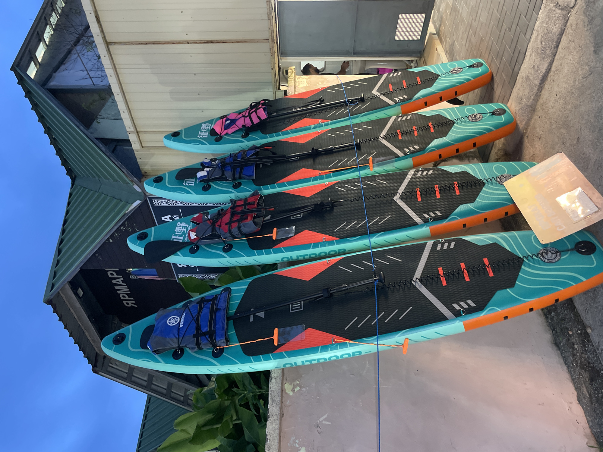 The image depicts a row of four stand-up paddleboards leaning against a textured, light-colored wall. The paddleboards are primarily teal with orange accents and black grip pads, and each has a life vest attached to its front. The paddleboards are arranged in a line, facing to the right, with their fins visible at the bottom. Behind the wall, a building with a green, corrugated metal roof is visible, and a sign with Cyrillic lettering, "ЯРМАРКА," is mounted on its dark facade. The sky above is a dusky blue, suggesting it is twilight. To the right of the paddleboards, a person, partially obscured, is visible inside a doorway or opening. The ground in front of the wall is paved with gray and light-colored rectangular stones. The scene is set outdoors, in what appears to be a commercial or recreational area.