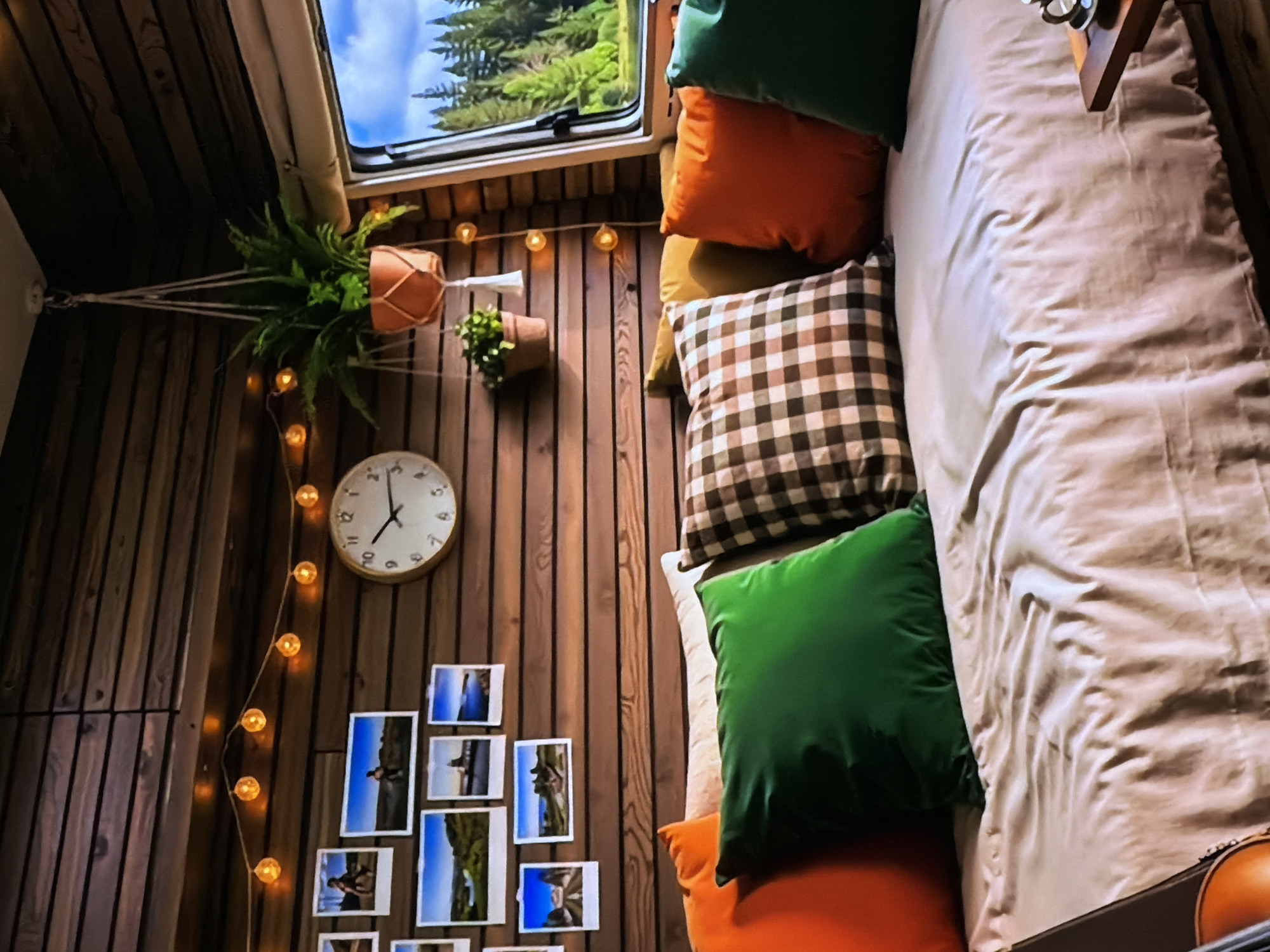 This image captures a cozy and rustic interior, likely of a camper van, RV, or a small cabin, situated in or near Sofiivska Borshchahivka, Ukraine. The dominant feature is a bed or daybed on the right side, adorned with several plush pillows in various colors including bright green, orange, and a brown and white plaid pattern, along with a light-colored duvet or blanket.
The walls and ceiling are paneled with warm-toned wooden planks, creating an inviting atmosphere. A rectangular window at the top left offers a glimpse of the outside, revealing a clear blue sky and lush green pine trees, suggesting a natural, possibly forested, setting during the daytime.
Decorative elements enhance the space's character: a hanging potted plant in a macrame hanger is visible on the left, and another small potted plant is mounted on the wall. A string of warm, globe-shaped fairy lights drapes vertically along the wall, providing ambient illumination, possibly for mood rather than primary light given the bright exterior. A round, analog clock with an off-white face and black hands is affixed to the wall, with the time appearing to be close to 12:50. Below the clock, a collection of small, possibly instant-print photographs or polaroids is arranged on the wall, depicting various outdoor scenes and possibly people, though individual details are indistinct.
No people are visible in the image, and no specific activity is taking place; the scene suggests a comfortable resting or living space designed for relaxation and enjoyment of nature. The overall impression is one of warmth, comfort, and a bohemian or adventurous spirit, ideal for travel or a tranquil retreat.