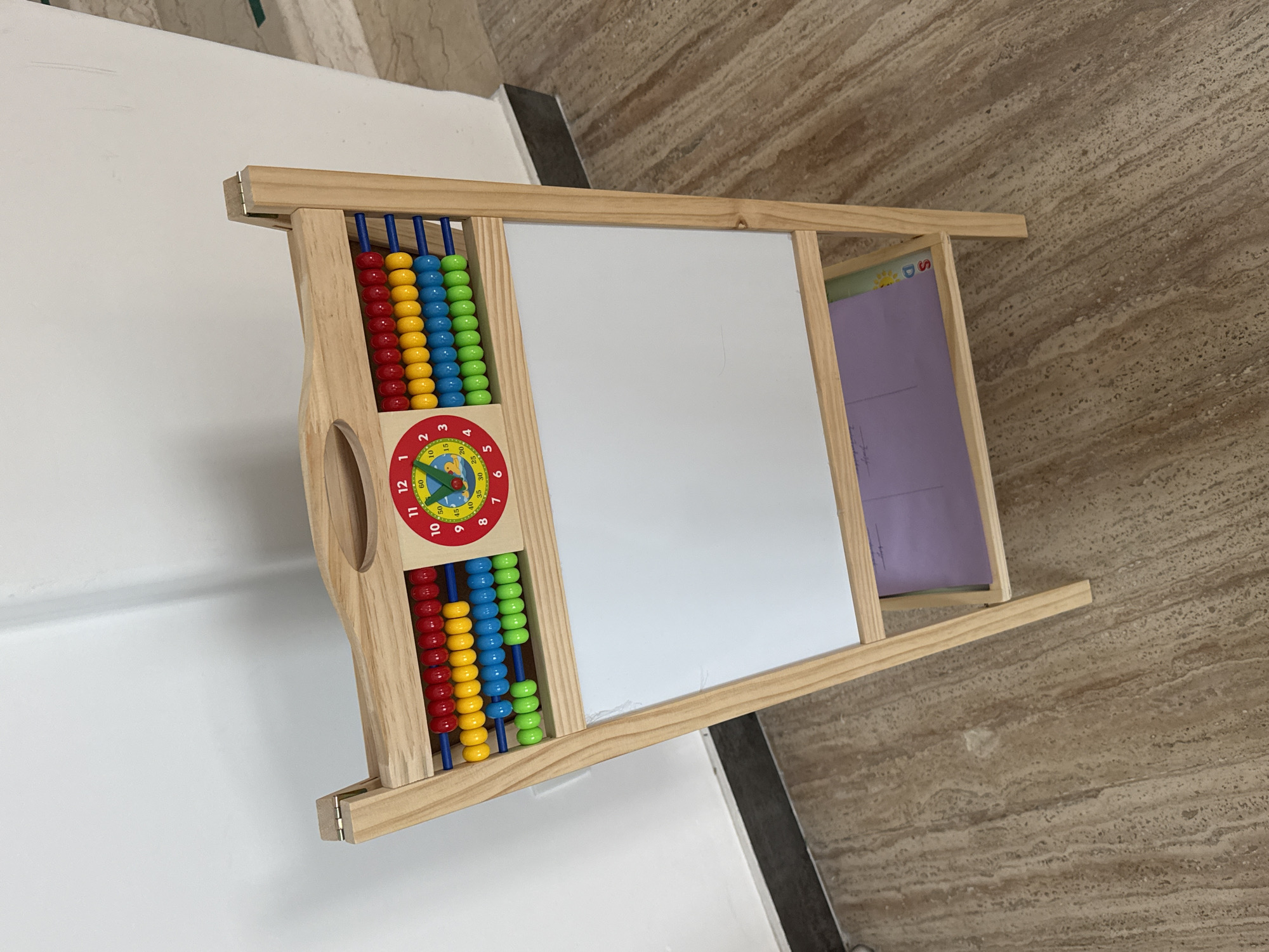 The media file captures a multi-functional wooden educational toy, likely for young children, placed indoors. The toy features a natural wood frame.
On the left side of the toy, an abacus is integrated, featuring four horizontal rods each holding ten colorful plastic beads. The beads are arranged in rows of red, yellow, blue, and green from top to bottom. Below the abacus, there is a circular clock face with numerical hour markers from 1 to 12, along with minute markings (5, 10, 15, 20, 25, 30, 35, 40, 45, 50, 60), and red and blue clock hands indicating time.
The central and largest part of the toy is a white dry-erase board, suitable for drawing or writing. To the right of the whiteboard, there's a smaller compartment where a light purple paper is inserted, partially visible. This paper appears to be a learning aid, with a partial sun graphic and the letter 'S' visible at its top right corner.
The background shows a plain white wall on the left and a light brown, patterned floor (resembling stone or large tiles) on the right. The lighting is bright and even, indicating an indoor daytime setting. No people or specific activities are depicted; the image is a still life of the educational toy. The image itself is rotated, making the toy appear on its side, though it is designed to stand upright.