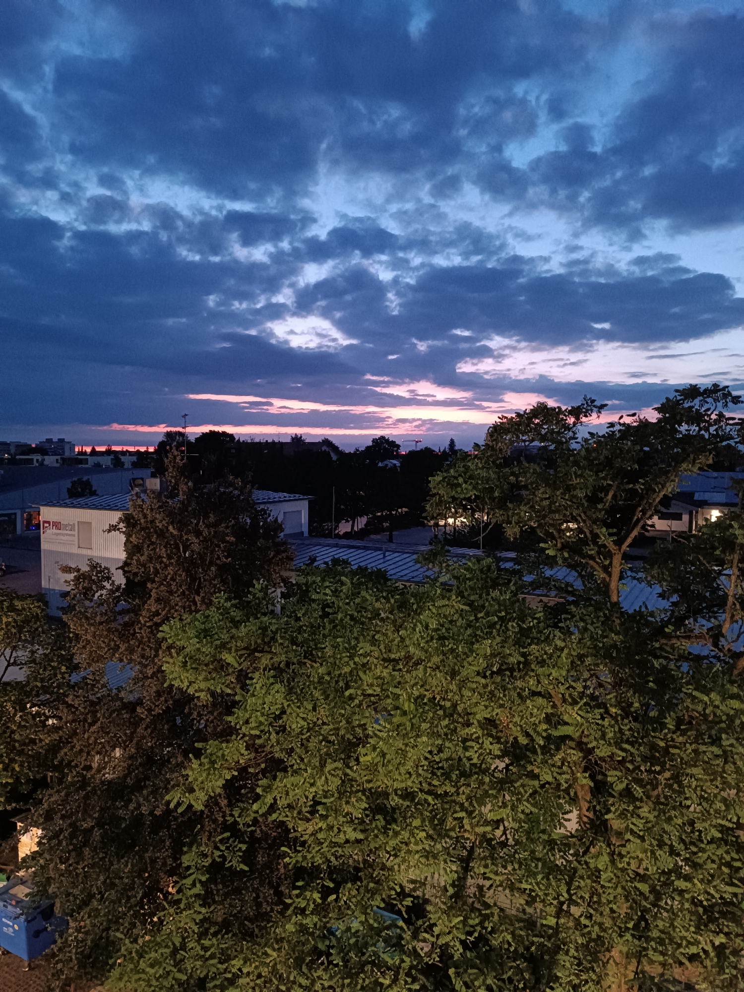 The image depicts a twilight sky over an urban landscape. The sky is a deep blue with scattered clouds tinged with pink and orange near the horizon, suggesting either sunrise or sunset. In the foreground, the dense foliage of trees obscures much of the lower portion of the scene.
Dominating the mid-ground is a white industrial building with a corrugated roof. A sign on the building reads "PRO metall". Beyond this building, more trees and rooftops of residential or commercial structures are visible. In the far distance, faint city lights and the silhouettes of buildings can be seen against the fading light. A crane is visible in the far left background, indicating ongoing construction.
To the left of the industrial building, at the bottom of the image, are two blue garbage bins. The overall impression is of an industrial or commercial area on the outskirts of a city, captured during the transition from day to night. The location is identified as Germering, Germany. There are no people visible in the image, and no discernible actions or interactions are taking place.
