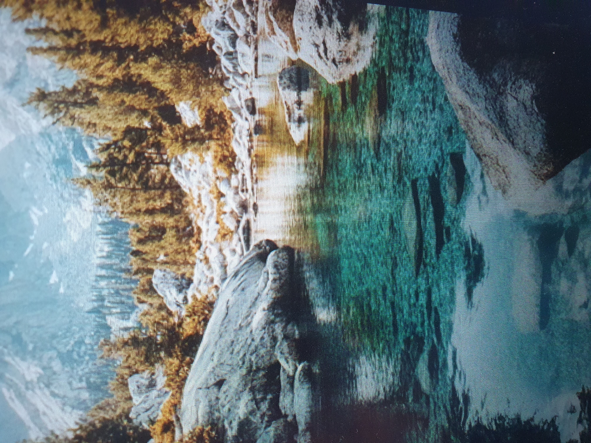 The image depicts a serene mountain landscape with a clear, turquoise body of water in the foreground. Large, grey rocks are scattered along the bank and in the water. The background features a dense forest of pine trees, transitioning into snow-capped mountains under a hazy sky. The scene appears to be a natural, undisturbed environment. There is no indication of any urban setting or any connection to the city of Tashkent, Uzbekistan.