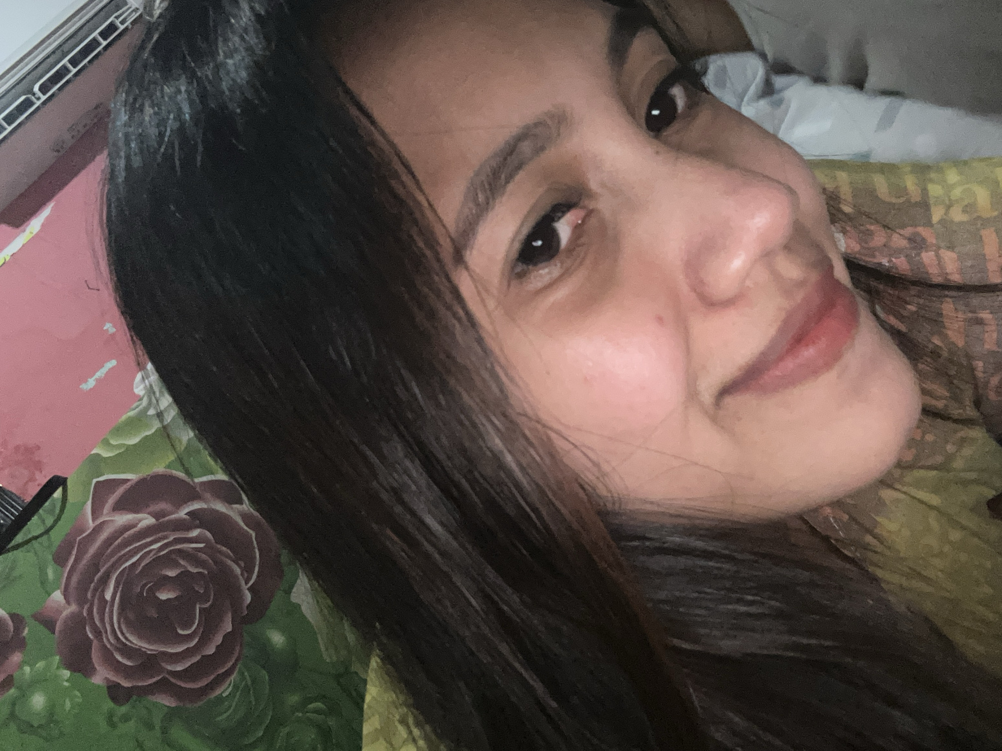A close-up selfie shows a young woman with long, dark hair and brown eyes smiling at the camera. She is wearing a patterned yellow shirt. Behind her, a wall displays a floral print with red roses and green leaves. The lighting suggests an indoor setting. The image does not provide enough detail to determine a specific location within Depok, Indonesia.