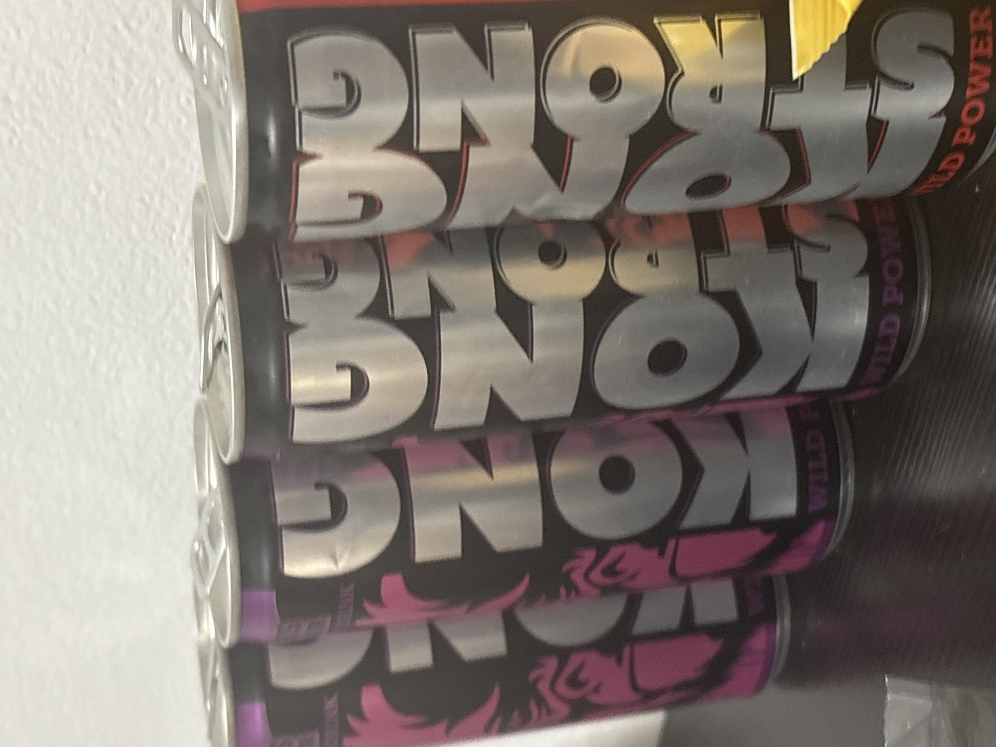 The image shows a row of energy drink cans on a dark surface. The cans are primarily silver with bold black and pink lettering that spells out "KONG". Some cans also have the words "WILD POWER" printed on them. The background is a white, textured wall. The lighting is somewhat dim, creating shadows on the surface. The scene is indoors, with no visible indication of its specific location within a city.