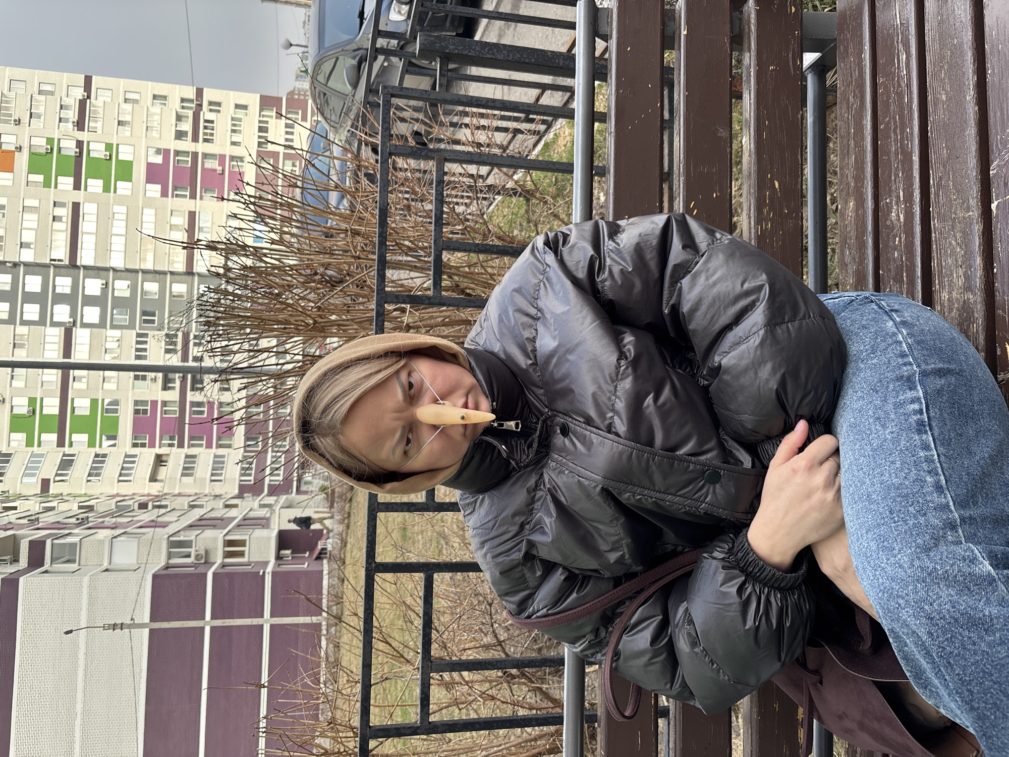 A person wearing a shiny black puffer jacket and blue jeans is sitting on a wooden bench. They also have a beige hat on and a long, wooden object attached to their nose with thin strings. The person is holding their hands together. Behind them is a black metal fence and some leafless bushes. In the background, there is a multi-story building with a distinctive pattern of white and colored blocks (green, purple, and grey). The scene appears to be located in Izhevsk, Russia, given the style of the building. There's a dark vehicle parked partially visible in the upper background. The sky is overcast.