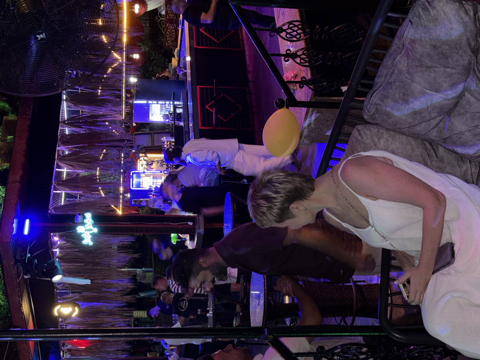 In Alanya, Turkey, an open-air bar with a thatched roof and vibrant neon lighting hosts a diverse group of people. A woman in a white dress is seated on a cushioned bench, holding a phone. To her right, a man with short blond hair and glasses is engaged in conversation with another man. In the background, a busy bar area is illuminated by blue and purple lights, with bartenders serving drinks. Several patrons stand at tables and the bar, some animatedly talking. A large industrial fan is mounted overhead. The atmosphere suggests a lively evening entertainment venue.