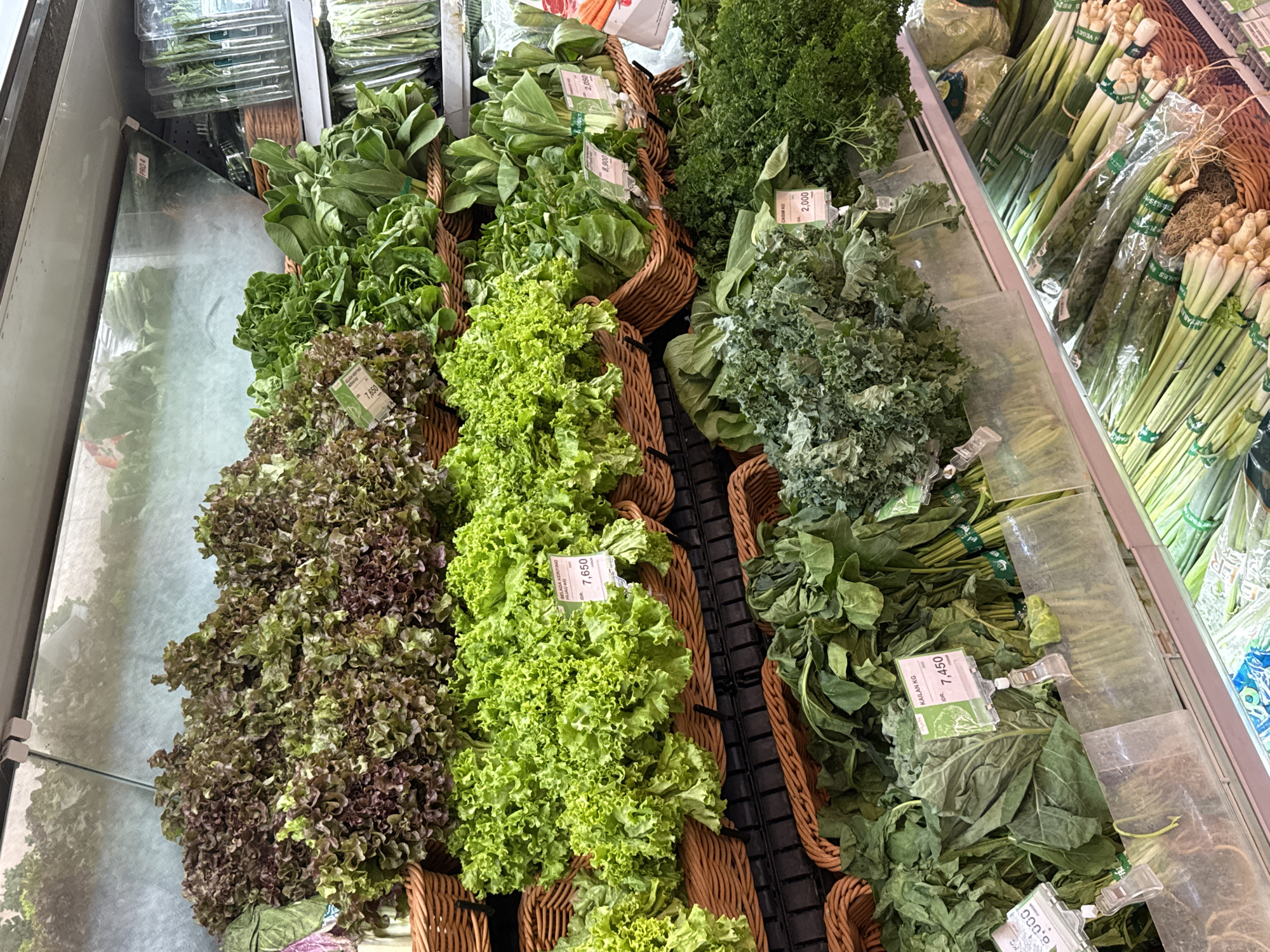 In a refrigerated display case at a grocery store in North Kuta, Indonesia, a variety of fresh leafy green vegetables are neatly arranged for sale. The display features multiple tiers filled with produce. The top tier showcases reddish-green lettuce and another type of leafy green. Below this, a large quantity of vibrant green leaf lettuce is presented in wicker baskets. The next tier down holds more leafy greens, including what appears to be bok choy and other varieties. The lower shelves display kale, parsley, and bundles of green onions and other long green vegetables. Price tags are visible in front of several of the produce sections, indicating prices in Indonesian Rupiah. The overall environment is clean and well-organized, typical of a modern supermarket produce section.