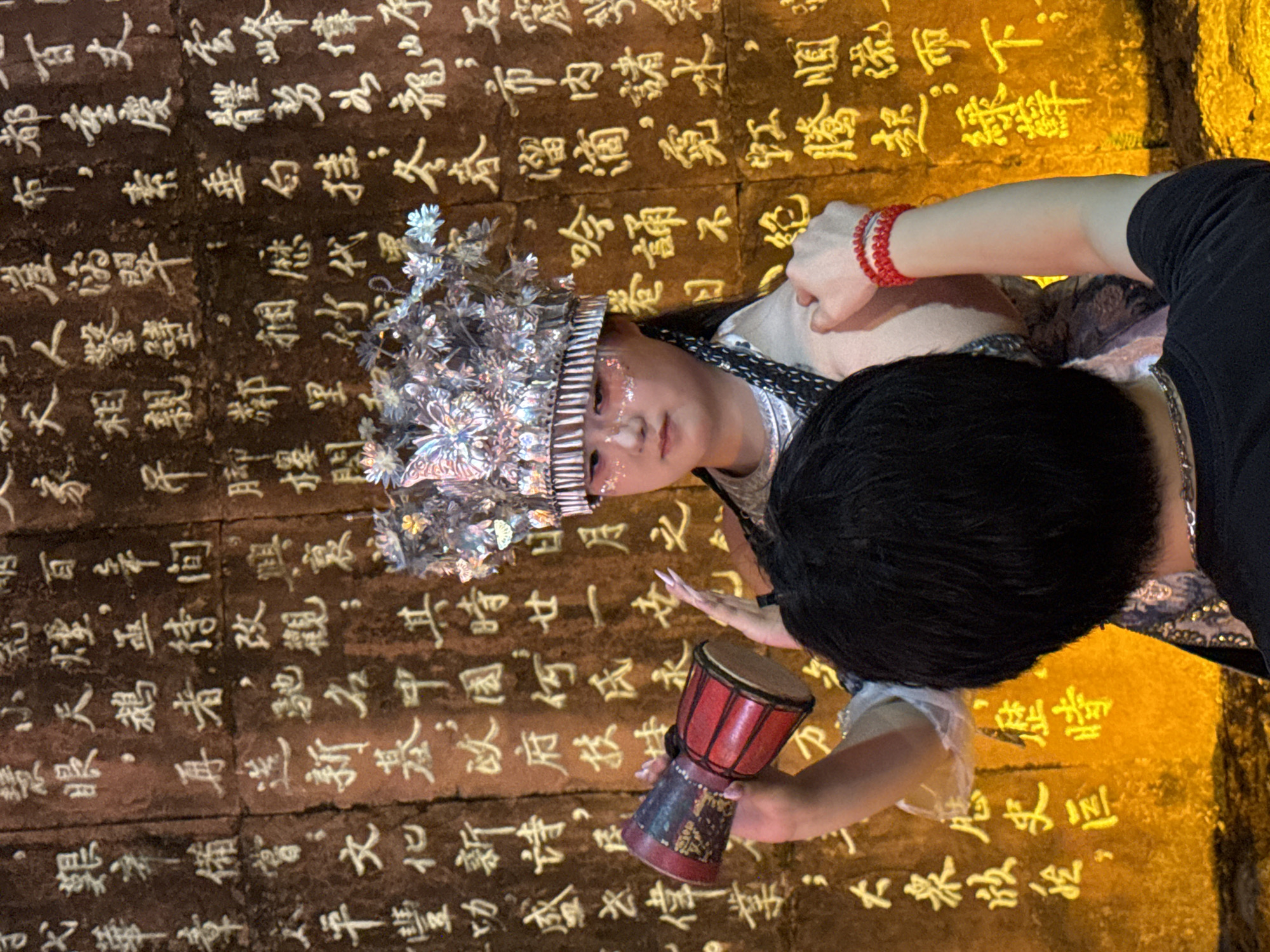 An image captured in Yuzhong District, China, shows a young woman adorned in a traditional headdress and makeup, holding a small drum. She is positioned in front of a wall inscribed with Chinese calligraphy. The woman is wearing a white garment with intricate patterns and a silver crown embellished with butterflies and floral motifs. Her face is painted with glitter and delicate lines, enhancing her theatrical appearance. The drum she holds is red and cylindrical, with decorative elements. Behind her, the wall's surface is covered with rows of Chinese characters, illuminated by a warm light that casts a glow on the scene. A person's arm, wearing a red beaded bracelet, is seen reaching towards the woman, suggesting an interaction or a gesture of assistance. The overall ambiance is evocative of a cultural performance or a historical reenactment, highlighting traditional Chinese artistry and performance.