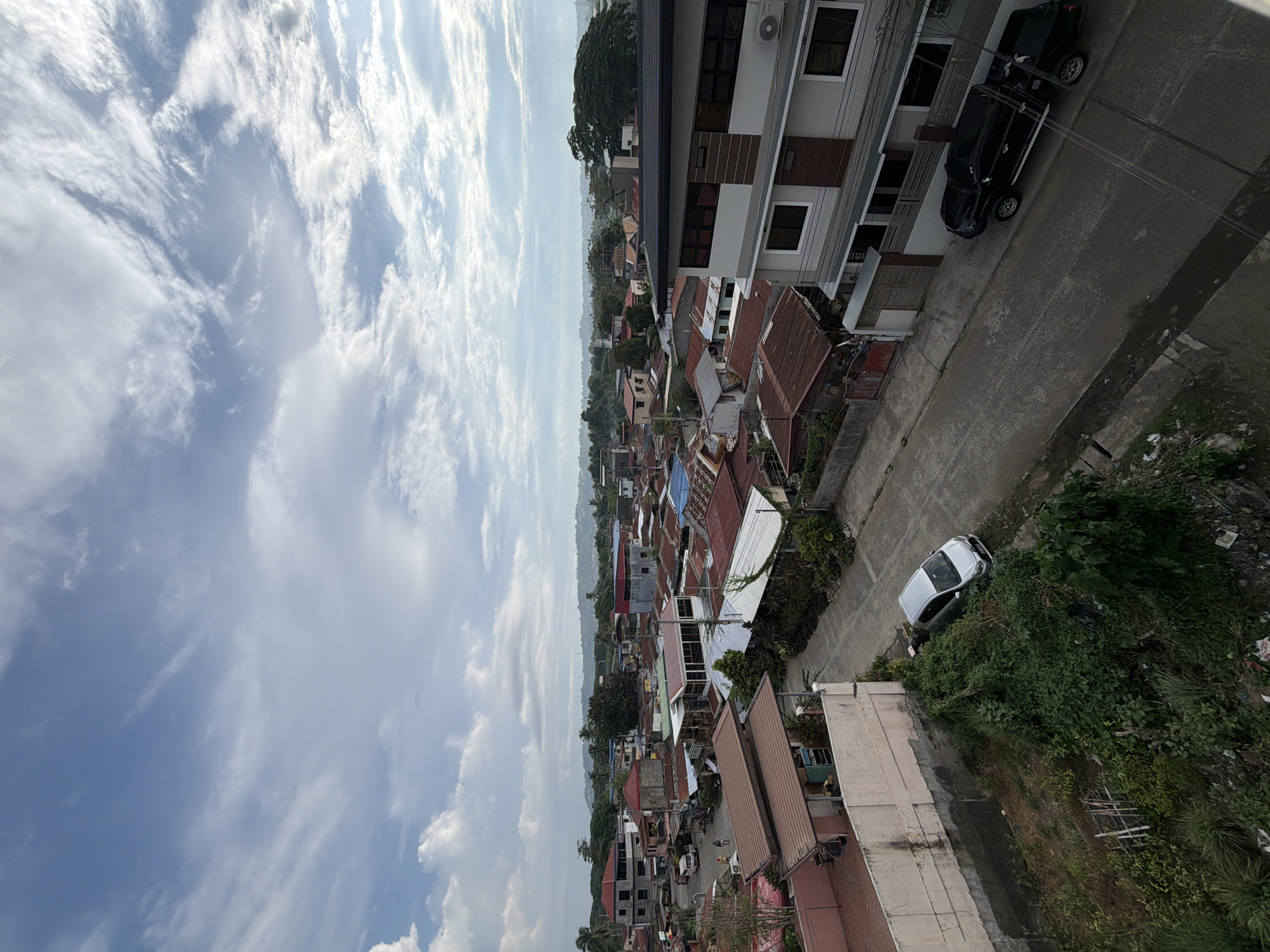 An aerial view shows a residential area in Davao City, Philippines. The sky, filled with scattered clouds, occupies the left side of the frame. A dense collection of houses with predominantly red and brown roofs extends into the distance. A two-story building is to the right, with a darker SUV parked on the road, with a newer white vehicle further down. Trees and greenery are interspersed among the houses and along the roadside. The road is concrete.
