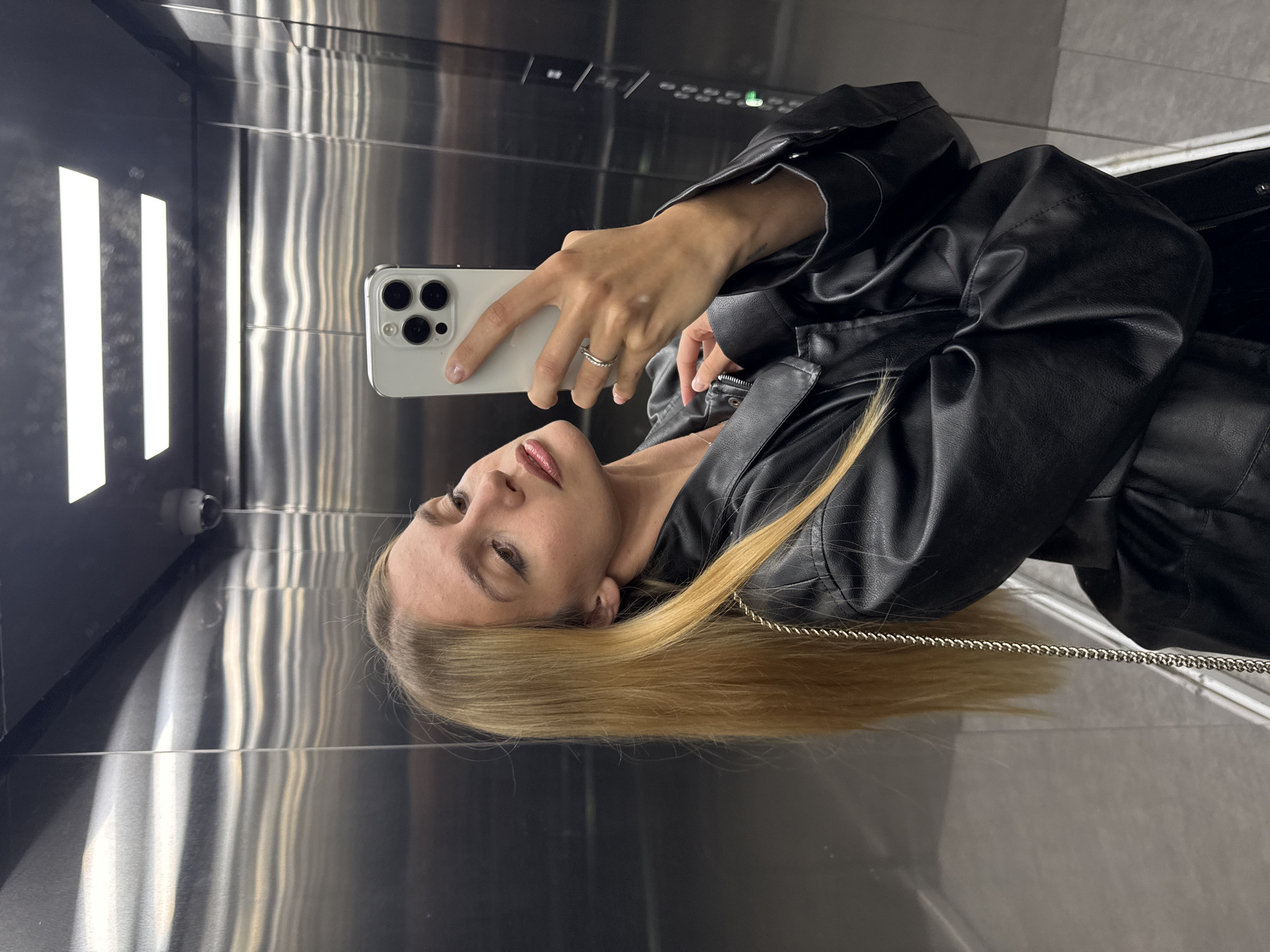 A young fair-skinned woman with blonde hair is taking a selfie in what appears to be an elevator. She is wearing a black leather jacket. The elevator is stainless steel with vertical striations. A security camera is visible in the top left corner of the image.
