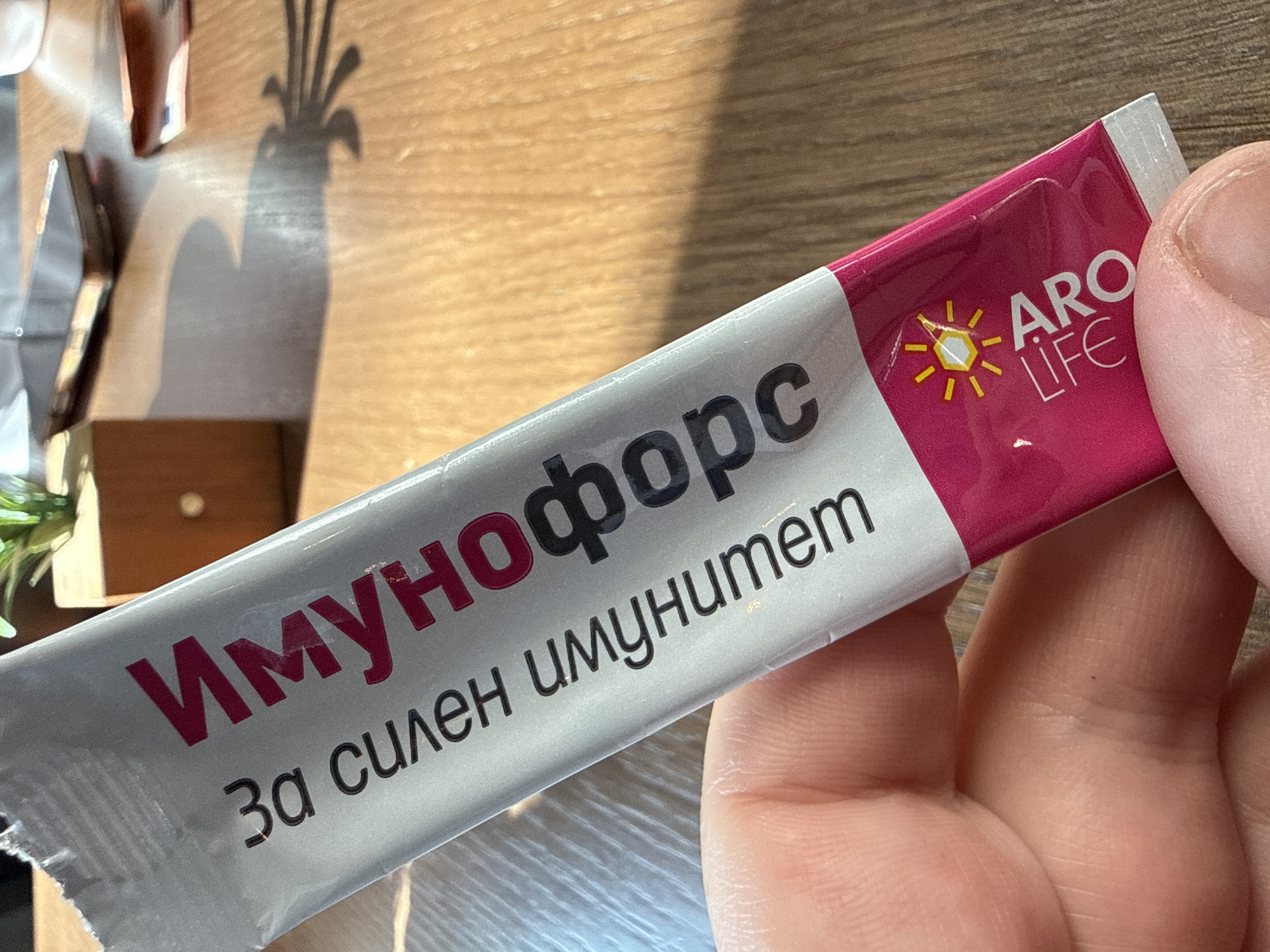 A human hand holds a silver and magenta sachet diagonally in the foreground. The sachet features the Cyrillic text "Имунофорс" (Immunoforce) in large magenta letters and "За силен имунитет" (For strong immunity) in smaller black letters. A yellow sun-like logo and the English text "ARO LIFE" are visible on the magenta portion. The background consists of a light-colored wooden table surface. On the table, a dark-colored smartphone lies flat, partially obscured. A small wooden cube pot containing a green plant is positioned behind the phone. Faintly visible to the right of the phone are partial views of what appear to be paper currency notes, potentially Euros. Strong directional lighting from the upper right casts distinct shadows of the plant and the sachet onto the table. This scene is located in Sofia, Bulgaria.