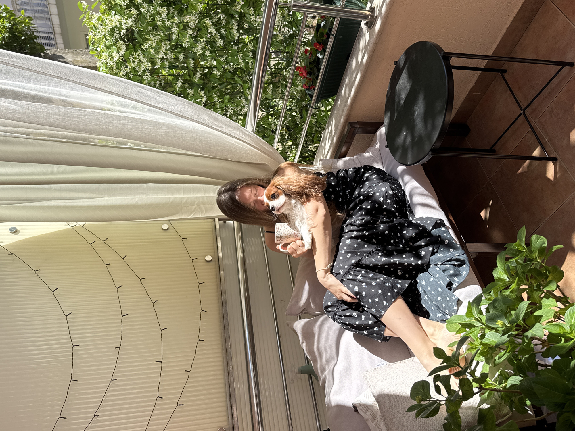 A woman is sitting with her dog on a cushioned bench on a balcony in Budva, Montenegro. The woman has long brown hair and is wearing a black dress with white stars. She is holding a Cavalier King Charles Spaniel in her arms. There is a black round table next to the bench, and a green plant is in the foreground. The balcony has a metal railing, and a sheer white curtain hangs to the left. Strings of lights are attached to a white wall behind the curtain. Green trees and foliage are visible beyond the railing. The floor of the balcony is tiled. The lighting suggests it is daytime and sunny.