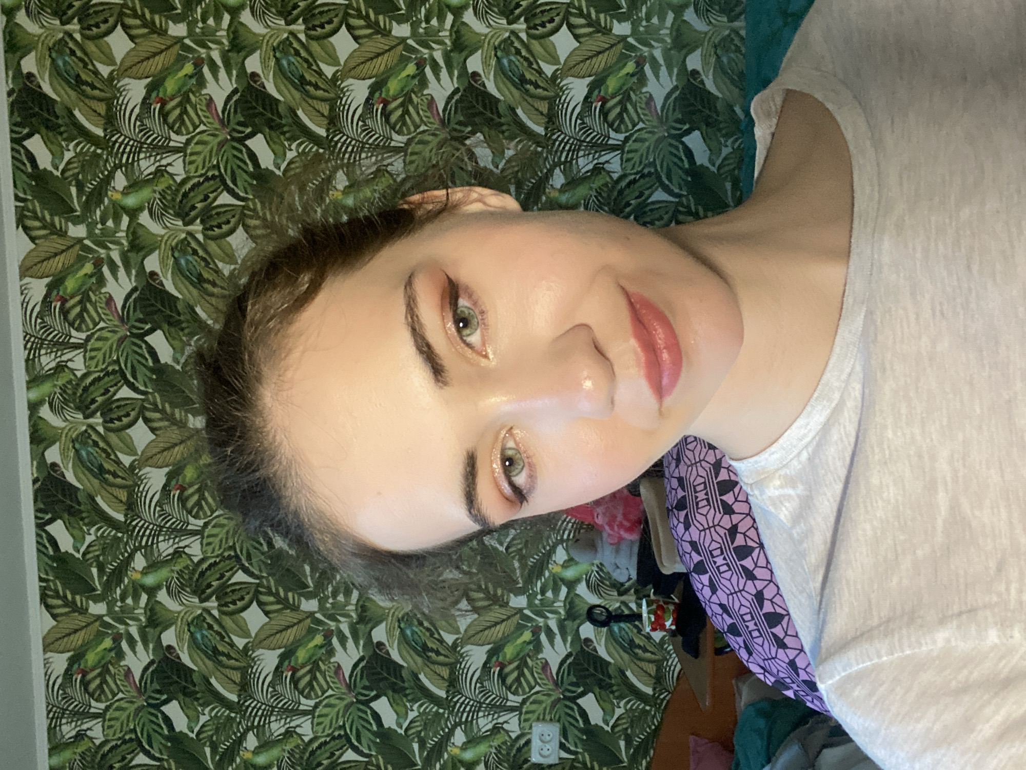 A young woman with light skin and green eyes is looking at the camera. She has on makeup including eyeshadow and lipstick. Her hair is dark brown and pulled up loosely. She is wearing a light-colored t-shirt. The background is a wall with a tropical-themed wallpaper featuring green leaves and parrots. To the left of the woman, there is an electrical outlet on the wall. Below that, a wooden table is visible with a mug and a black object. Behind the woman, a bed or sofa is partially visible with a purple and black patterned blanket. To the far left, a dark green blanket is also visible. The scene appears to be indoors, likely in a residential setting. The image does not contain any elements that would allow for the identification of the city of Zavodoukovsk, Russia.