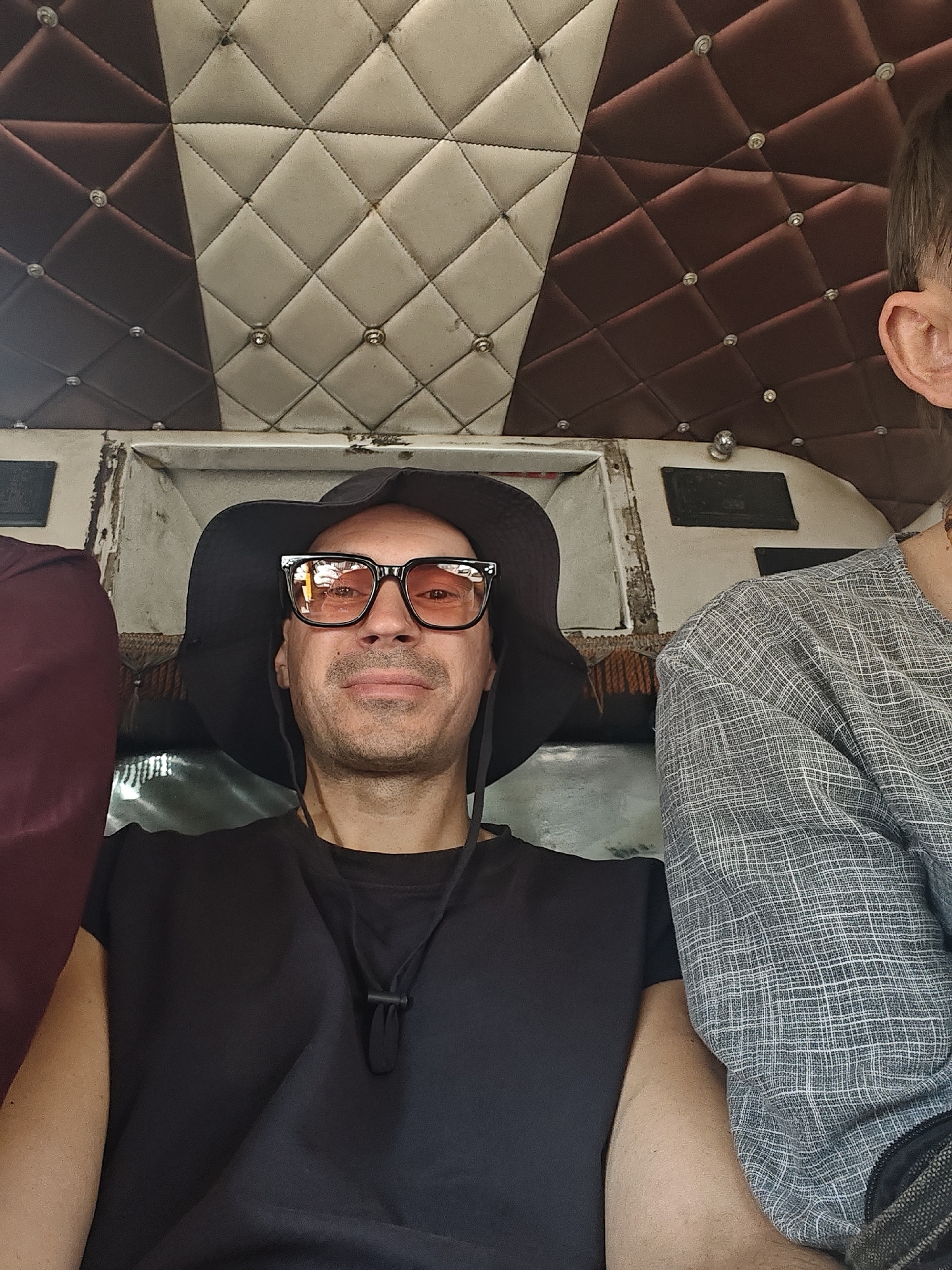The image captures three individuals seated within the interior of a vehicle in Unknown, Unknown. The primary subject, an adult male, occupies the central foreground, looking directly at the camera with a neutral expression. He wears a black wide-brimmed bucket hat, black-framed sunglasses with amber-tinted lenses, and a plain black t-shirt with an adjustable black cord around his neck. To the left of the central subject, a person's arm and shoulder, clad in a dark red/maroon garment, are partially visible. To the right, the right ear and upper torso of another individual are visible; this person wears a light grey long-sleeved shirt with a subtle grid pattern. The vehicle's ceiling is upholstered with a diamond-quilted pattern, divided into an off-white section on the right and a dark brown section on the left, each diamond intersection featuring a metallic stud. Below the ceiling upholstery, a worn and discolored light-colored structural beam or trim is present, with additional damaged interior paneling visible beneath.