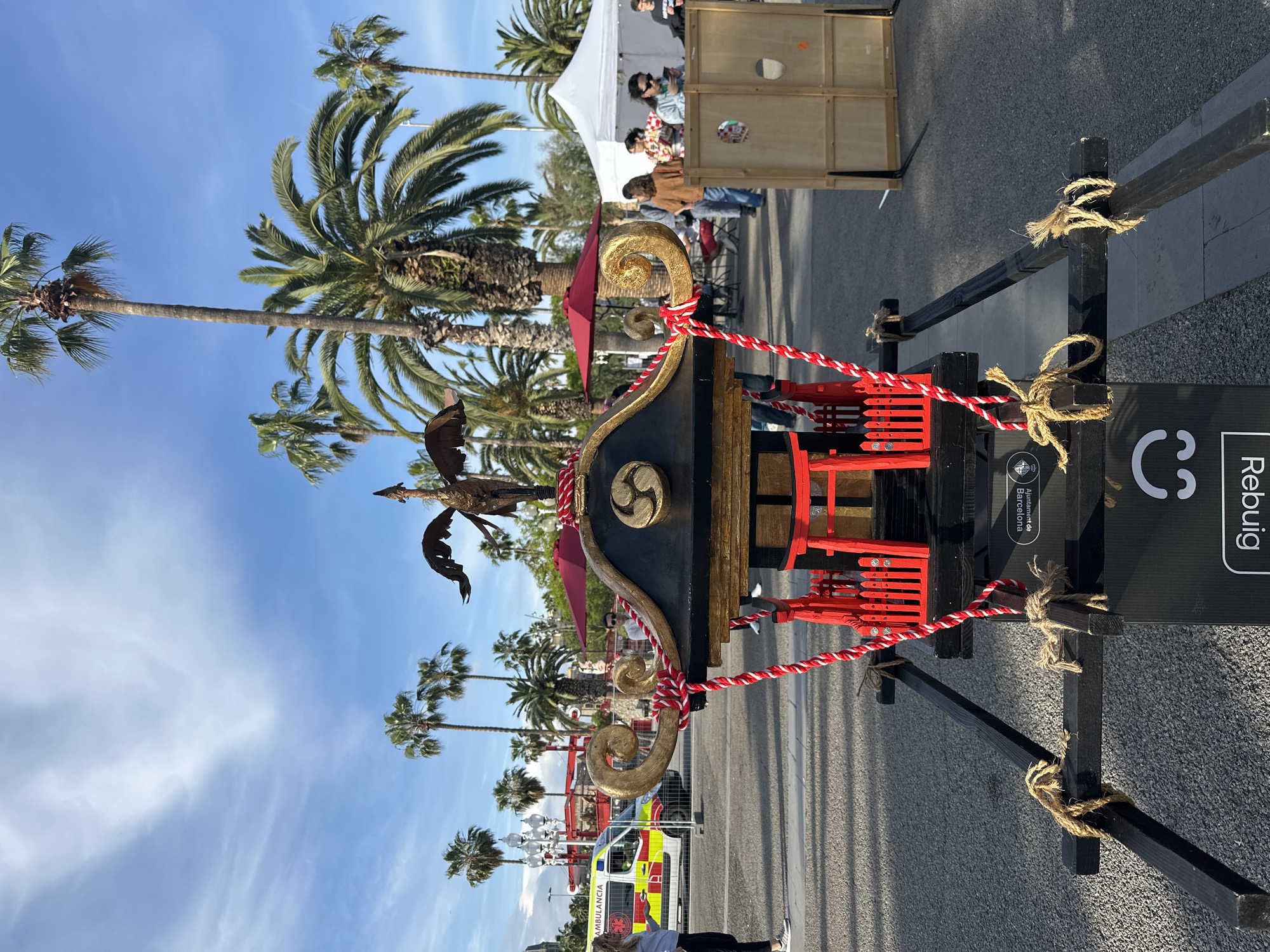 The image shows a Japanese-style portable shrine, or Mikoshi, situated outdoors on a paved surface. The Mikoshi is black, gold, and red, with ornamental ropes tied around it. Palm trees are visible in the background against a blue sky with scattered clouds.
Behind the Mikoshi, there is an ambulance and a vendor tent. People are visible near the tent and further away on the street.
A sign with the word "Rebuig" and a "C" symbol is attached to the Mikoshi stand. The sign also includes the text "Ajuntament de Barcelona," suggesting that the location is in Barcelona, Spain.