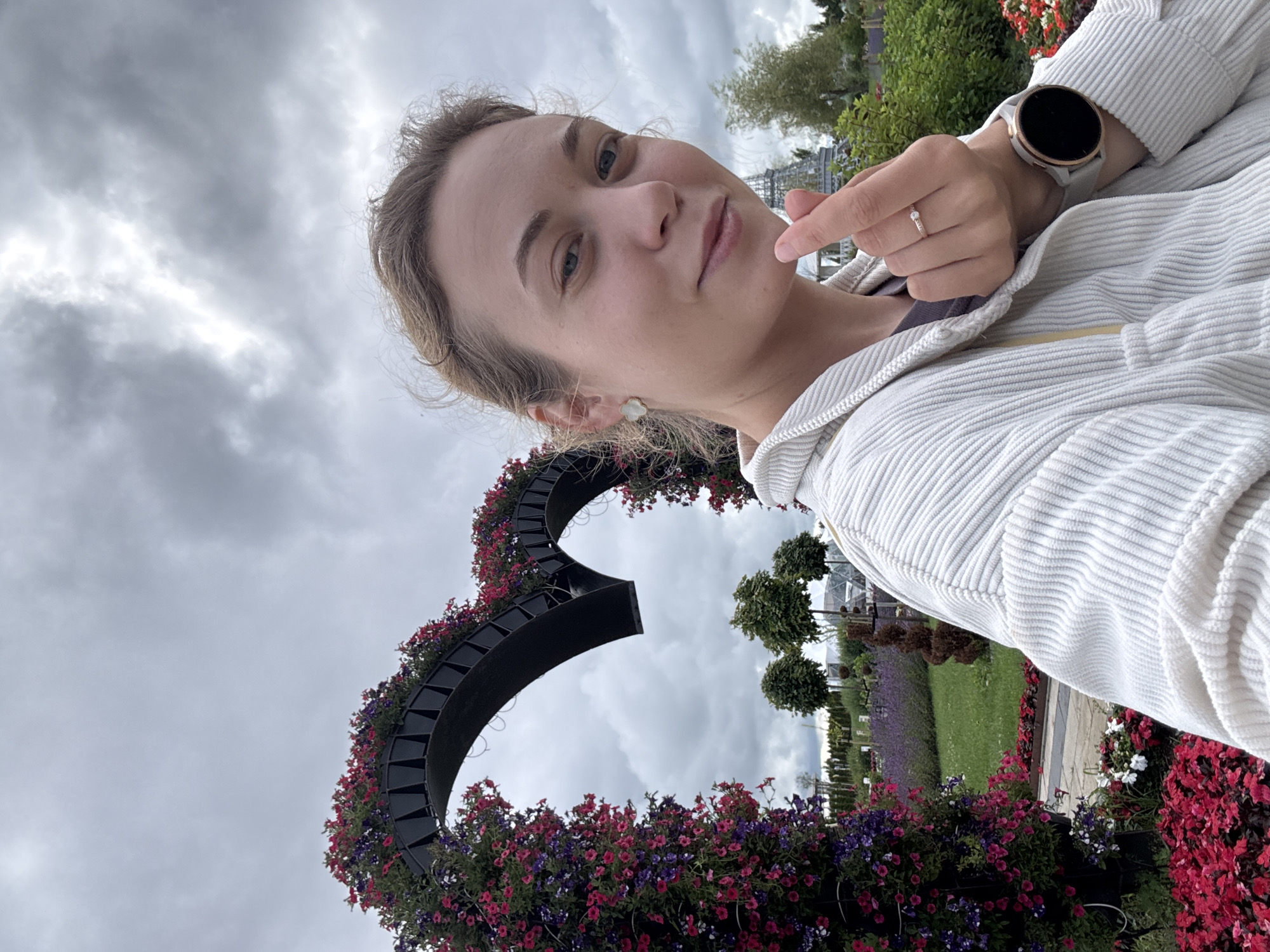 A young woman with blonde hair pulled back is taking a selfie outdoors in a garden. She is wearing a white corduroy jacket. Her left hand is raised to her face, forming a finger heart gesture. She is wearing a ring on her left ring finger and a smartwatch on her left wrist. Behind her, a large heart-shaped arch is covered in pink and purple flowers. The background shows a park with trees, manicured bushes, and a cloudy sky.