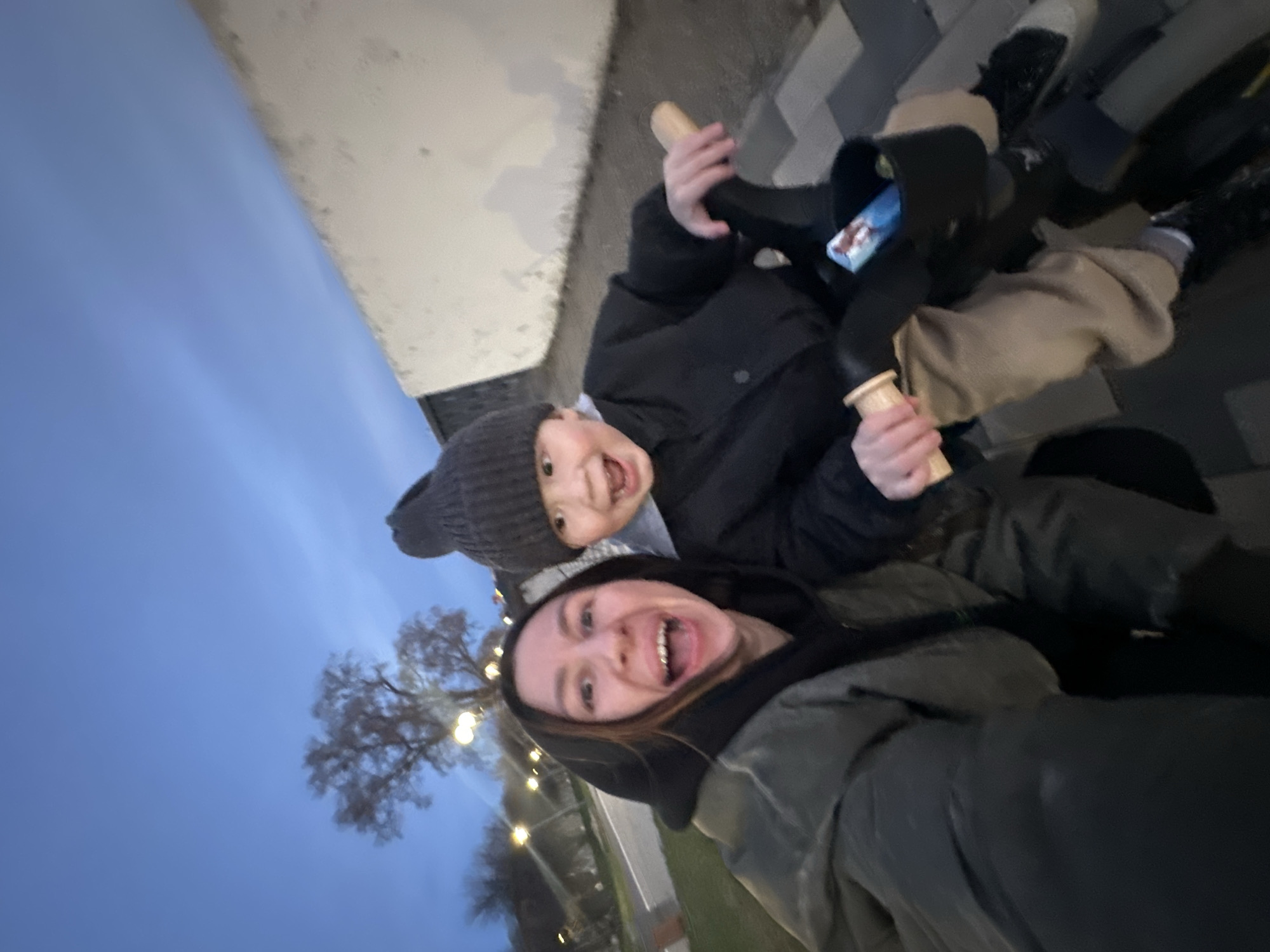 A joyful, tilted selfie captures an adult and a child outdoors in Votkinsk, Russia, during what appears to be dusk or night. The sky is a deep blue, with blurred decorative lights visible in the background, suggesting a festive or atmospheric setting.
In the foreground, a young adult, likely a female, smiles broadly at the camera, her mouth open in laughter or excitement. She has dark hair and is wearing a dark hooded jacket, possibly with a grey or olive-green outer layer. Slightly behind and above her, a young child also grins widely with an open mouth, showing teeth. The child is dressed warmly in a dark grey knitted beanie hat and a thick black winter jacket, indicating cold weather.
The child holds two toy-like objects that resemble ice cream cones, featuring light wooden handles and dark, rounded tops. One of these toys has a small, rectangular blue and white packet attached, though any text on it is illegible due to blurriness.
The background reveals bare trees, consistent with a colder season like late autumn or winter. Blurry circular lights, possibly from string lights, are visible amongst the trees. The ground below the subjects is a patterned grey and black paved surface, suggesting a sidewalk or public square. A light-colored wall or structure runs along the upper edge of the frame. Both individuals express strong positive emotions, conveying a sense of happiness and shared amusement during their outdoor activity.