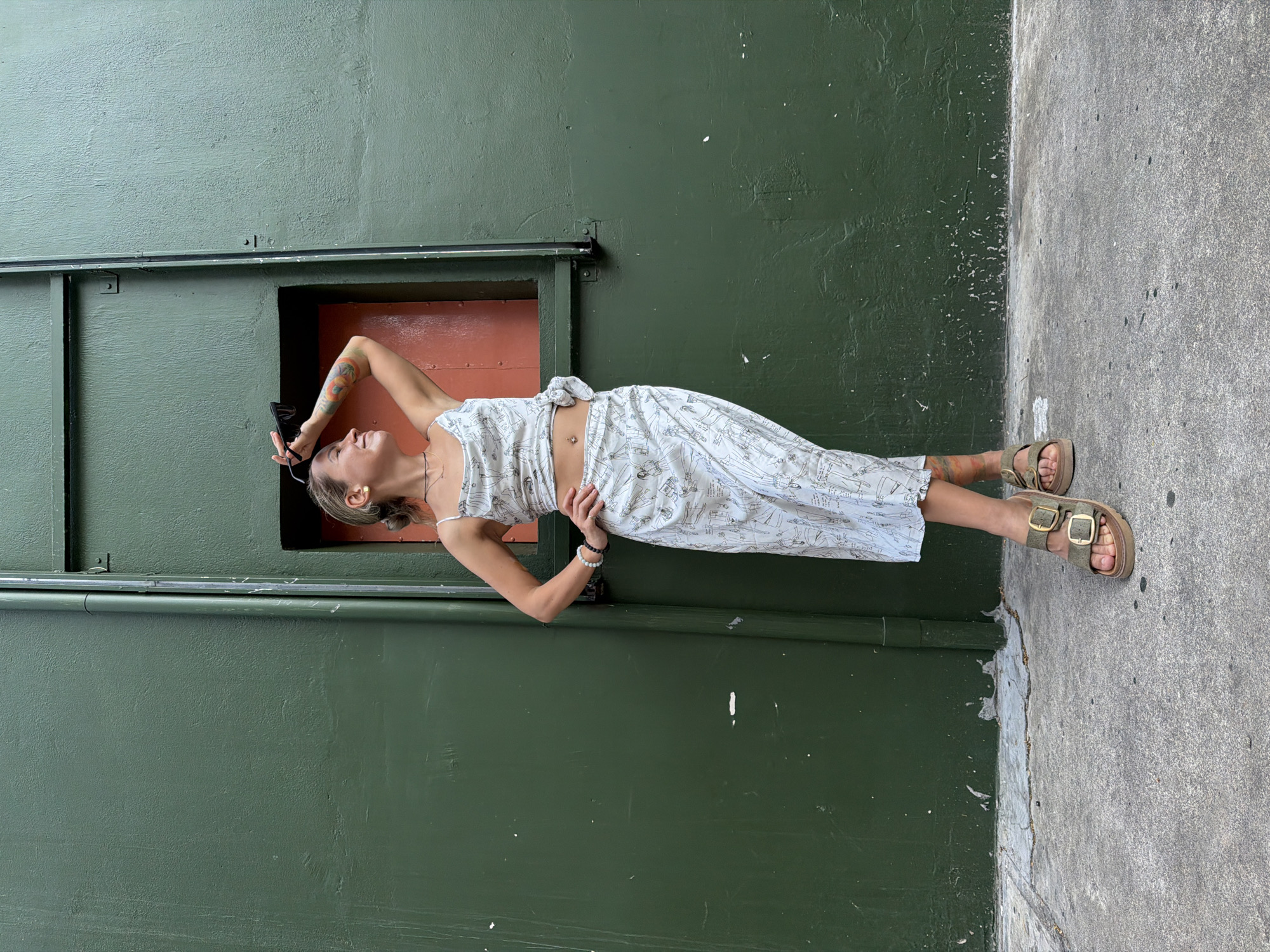 A young woman stands in front of a dark green wall with a narrow orange opening. She is wearing a white two-piece outfit with a black line-art pattern. The top is a cropped camisole with a knot in the front, and the skirt is a mid-length, flowy style. She has her right arm raised, holding sunglasses, and her left hand on her hip. The woman has tattoos on her arms and legs. She is wearing chunky, olive-green sandals. The ground in front of the wall is a gray concrete sidewalk. The wall appears to be part of an urban building.