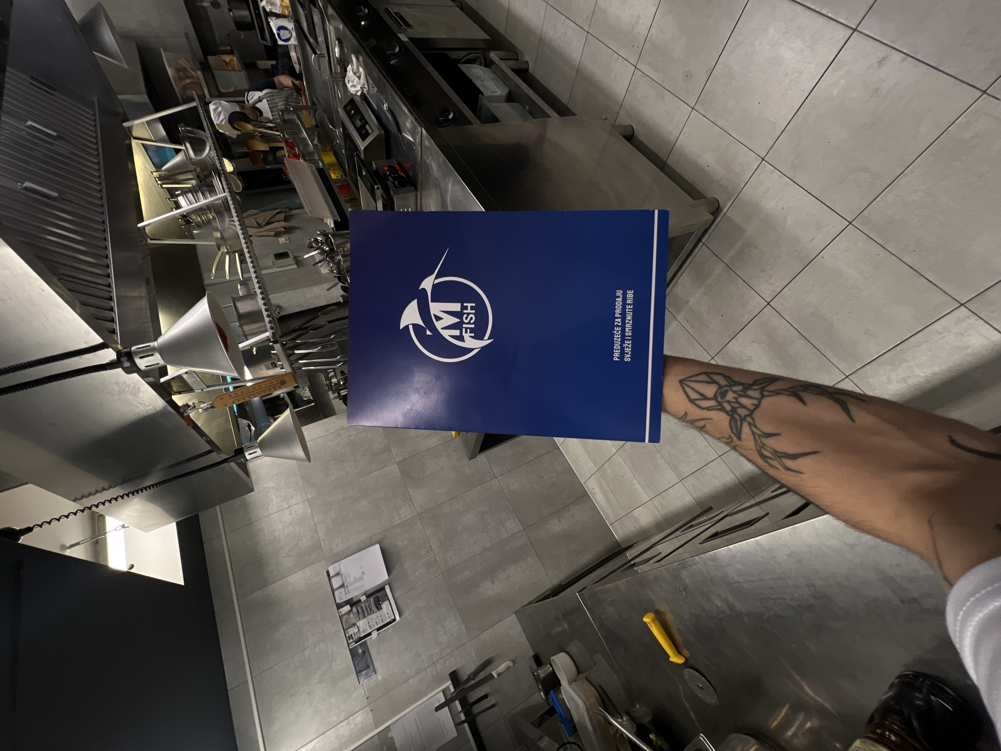 A medium shot from a high-angle perspective shows a commercial kitchen with stainless steel counters and equipment. Predominantly neutral gray tiles cover the floor and portions of the walls.
The central subject is a navy blue folder with a white logo reading "M FISH" and accompanying text in a local language, held by a tattooed arm extending into the frame from the lower right. The tattoo features stylized black ink artwork.
In the background, commercial cooking equipment and kitchen utensils are visible. A cook is seen working at the back.
A closer inspection of the floor reveals a laminated sheet with several miniature diagrams.
While the exact location in Budva, Montenegro, cannot be definitively determined from the image, the presence of the M FISH folder suggests the kitchen might be affiliated with or supplying a restaurant or business operating under that brand.