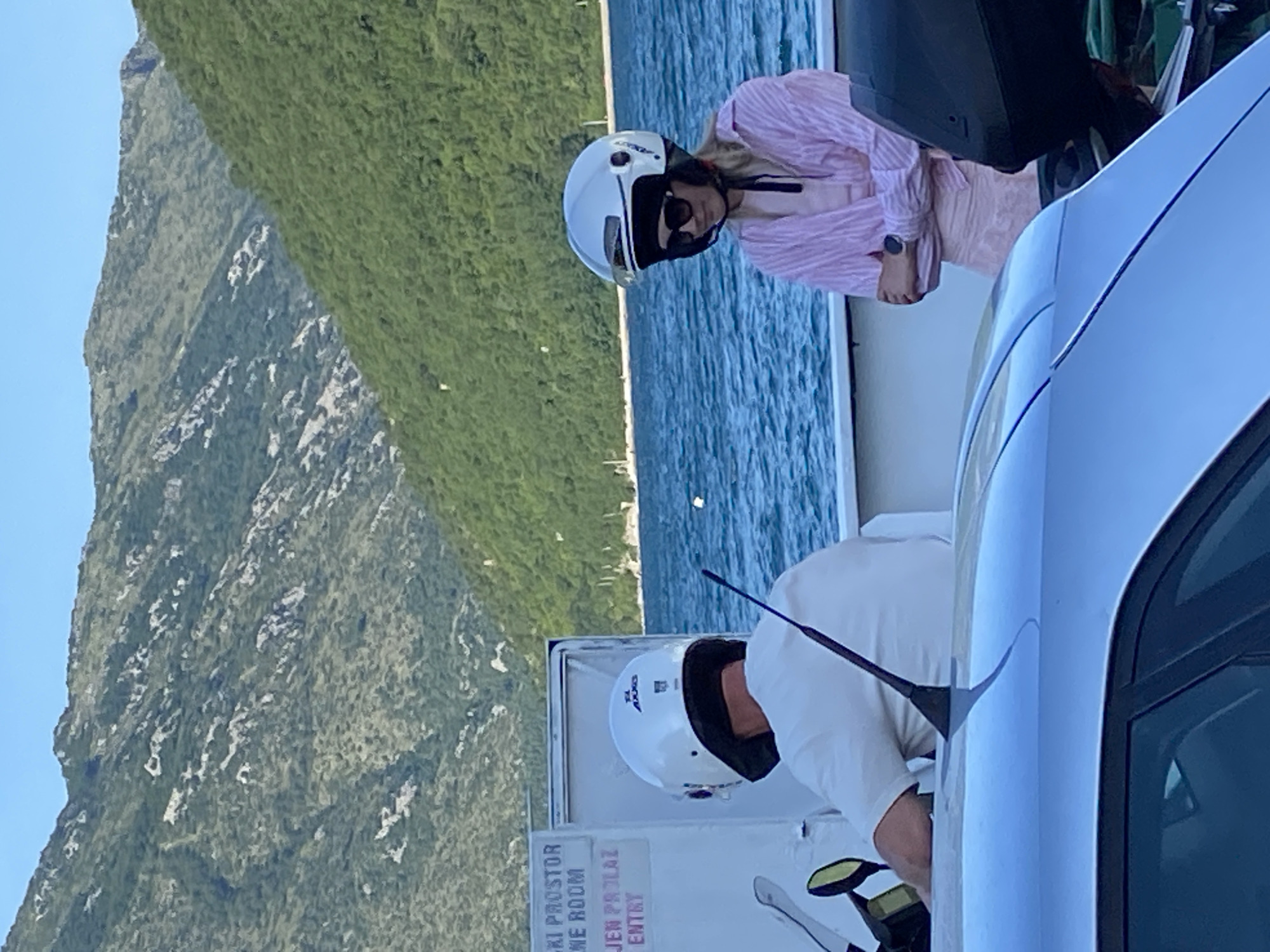 Captured in Đurići, Montenegro, the image shows a mountainous backdrop and a stretch of water. A woman wearing a white helmet, sunglasses, and a pink-striped shirt stands near a person in a white shirt and white helmet, suggesting they may be involved in an activity related to personal transportation. Part of a white car is visible in the frame, adding to the scene's context. There is a sign that says "SKI PROSTOR, SVLACIONE ROOM, JEN PROLAZ, ENTRY."