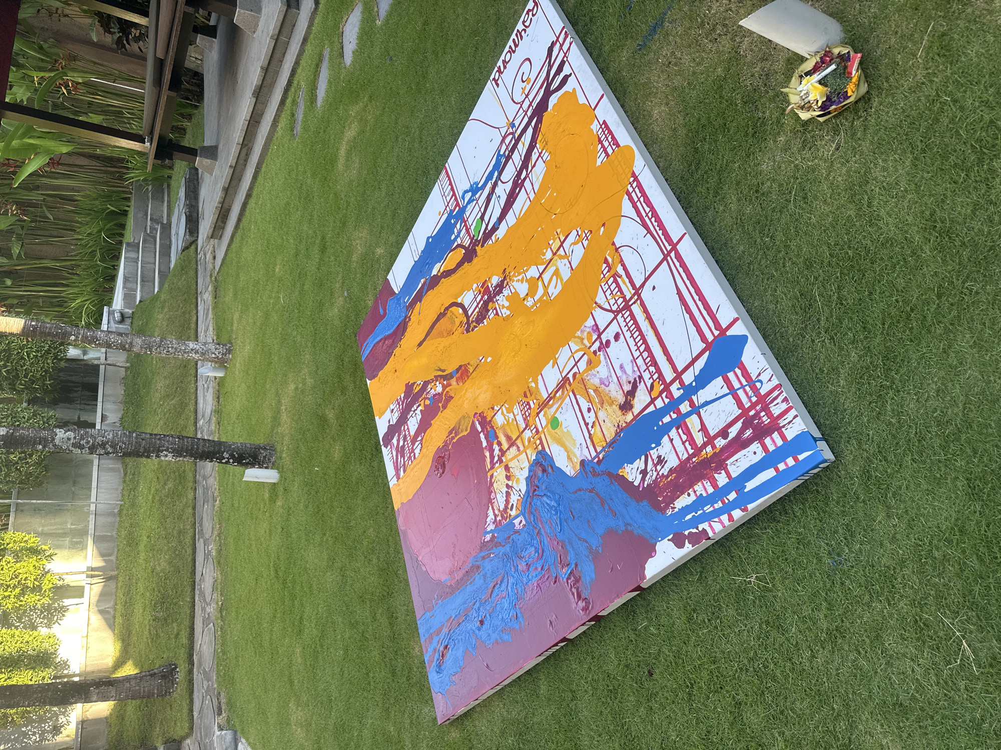 The image shows a large, abstract painting resting on a well-maintained lawn in North Kuta, Indonesia. The painting features a white background with a grid-like pattern and vibrant splashes of orange, blue, and red paint. The painting is titled "Raymond" along its edge. The environment includes lush green grass, a nearby building with a stepped entrance, and a few trees, including a palm tree. A small, woven basket containing flowers and leaves sits on the grass near the painting. The overall scene appears to be outdoors, potentially in a garden or yard.