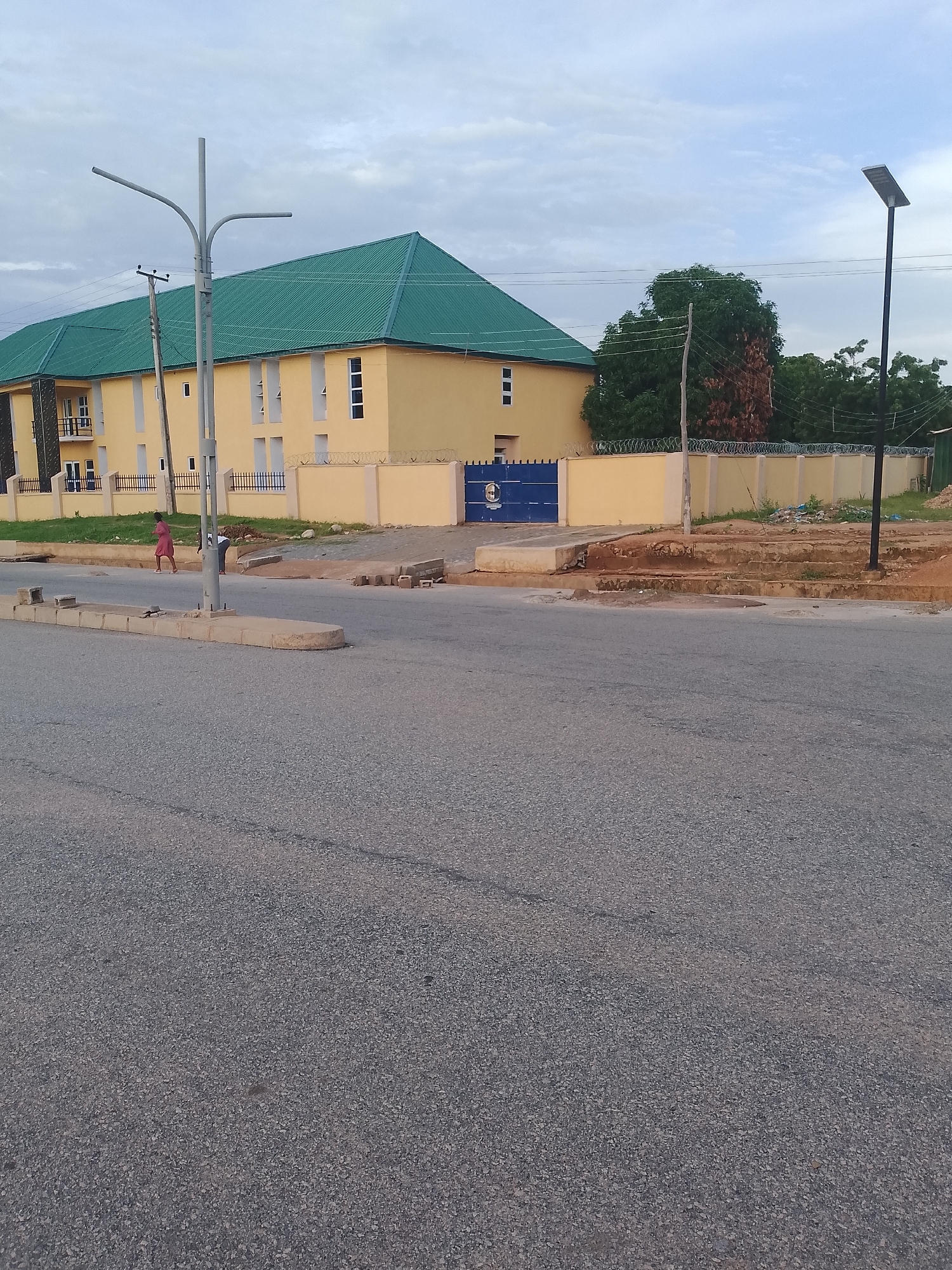 The image depicts a wide street in front of a two-story yellow building with a green roof. The building has several windows and a blue gate with a circular emblem. A street lamp stands on the left side of the image, and a solar-powered street lamp is visible on the right. A few people are present near the building, one in a pink dress and another bent over. The foreground is dominated by the asphalt road, with concrete curbs and some debris along the side. Trees and a cloudy sky are visible in the background.