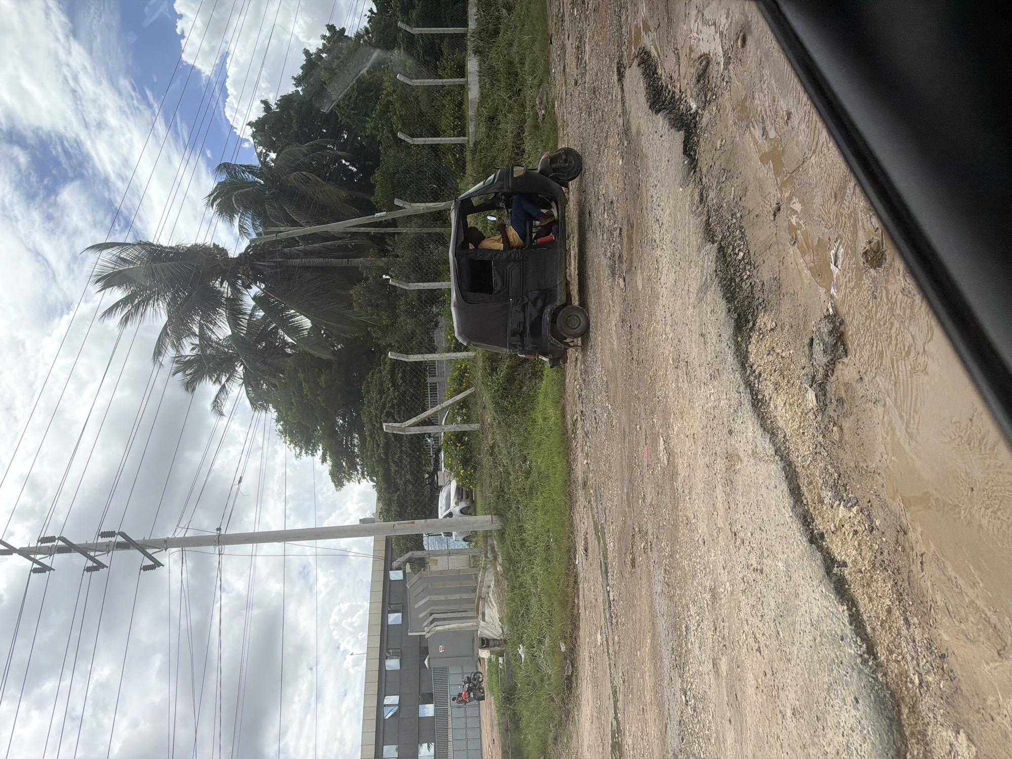 A muddy, deeply rutted dirt road is the primary focus of the image, with a three-wheeled vehicle (tuk-tuk) visible on the road. The tuk-tuk is black with a yellow-clad person inside. A building with multiple windows is seen on the left side of the road, and a motorcycle with a rider can be seen near the building. The background features palm trees, other vegetation, a chain-link fence, and power lines against a sky with scattered clouds. The photograph was taken from inside another vehicle. The location of the city and area are unknown.